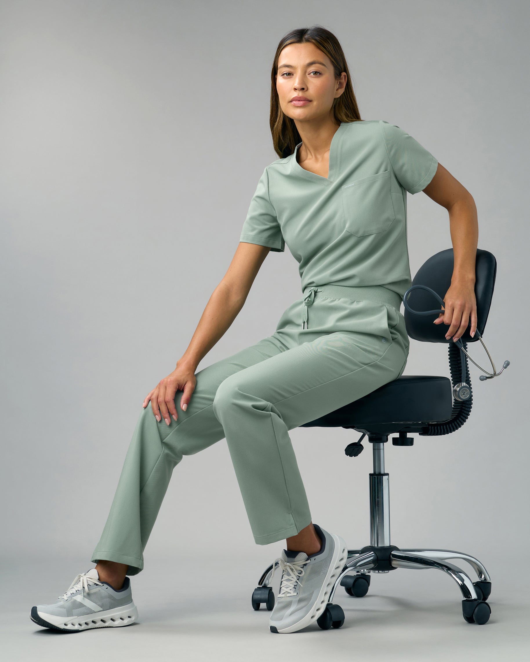 Woman in a sage green scrub top and matching knit-waist scrub pants sitting on a rolling stool, holding a stethoscope