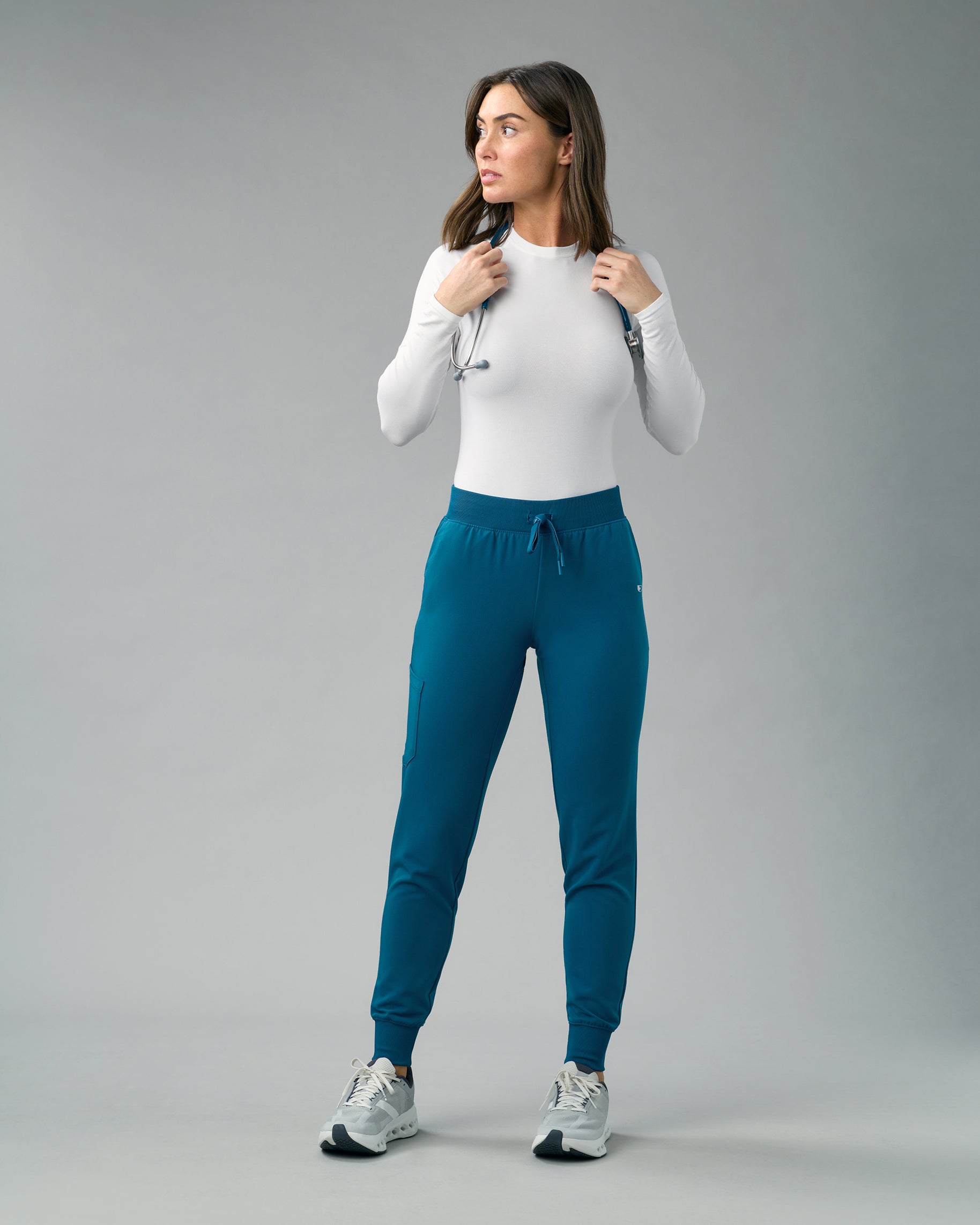 Woman in a white long-sleeve top and teal slim scrub joggers with a stethoscope around her neck