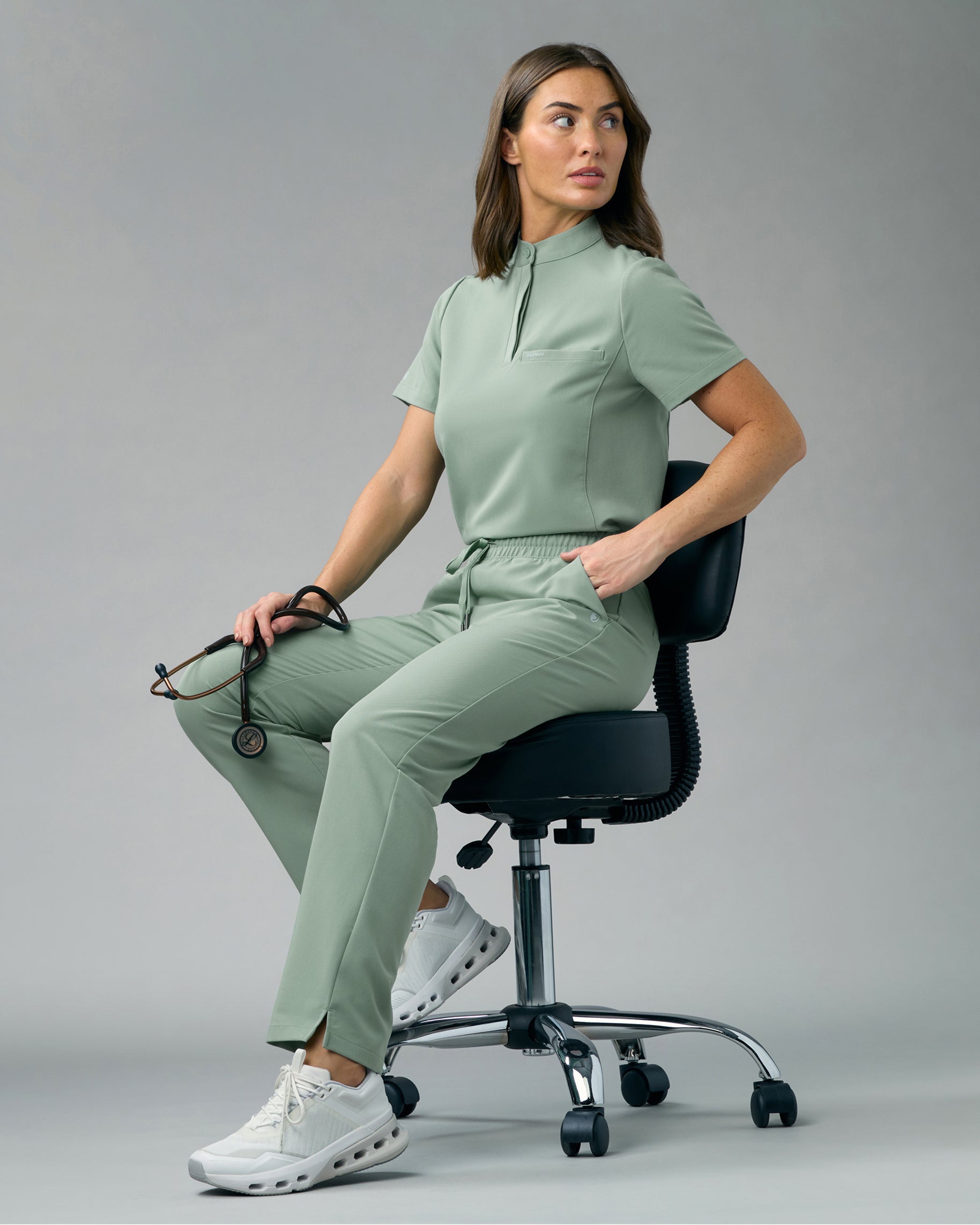 Woman in a sage green stand-collar scrub top and high-waisted scrub pants sitting on a rolling stool, holding a stethoscope