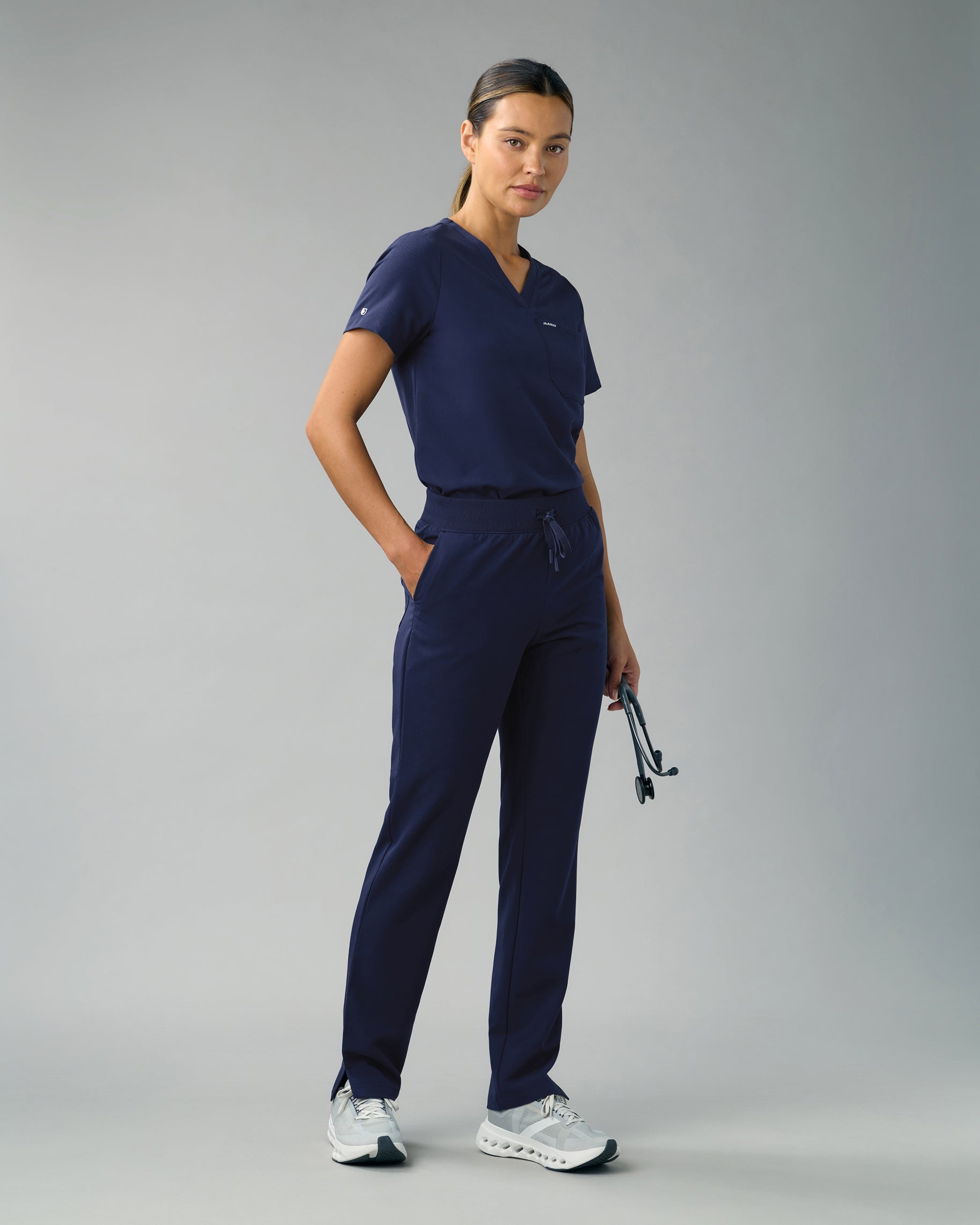 Woman in a navy V-neck scrub top and slim knit-waist scrub pants with one hand in her pocket and a stethoscope in the other