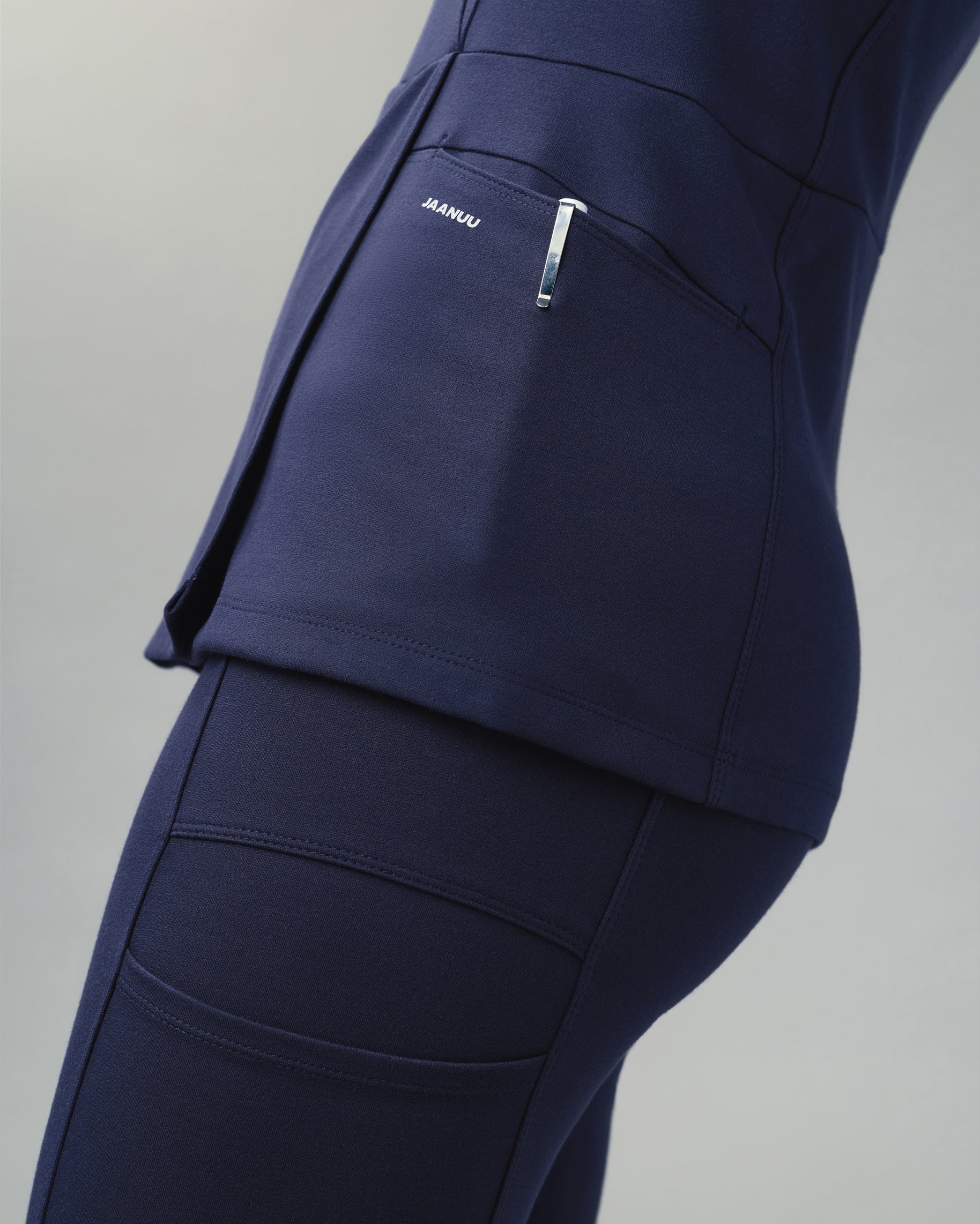 Side view of a navy mock-wrap scrub top with a pen clipped in the side pocket marked JAANUU, over matching scrub pants