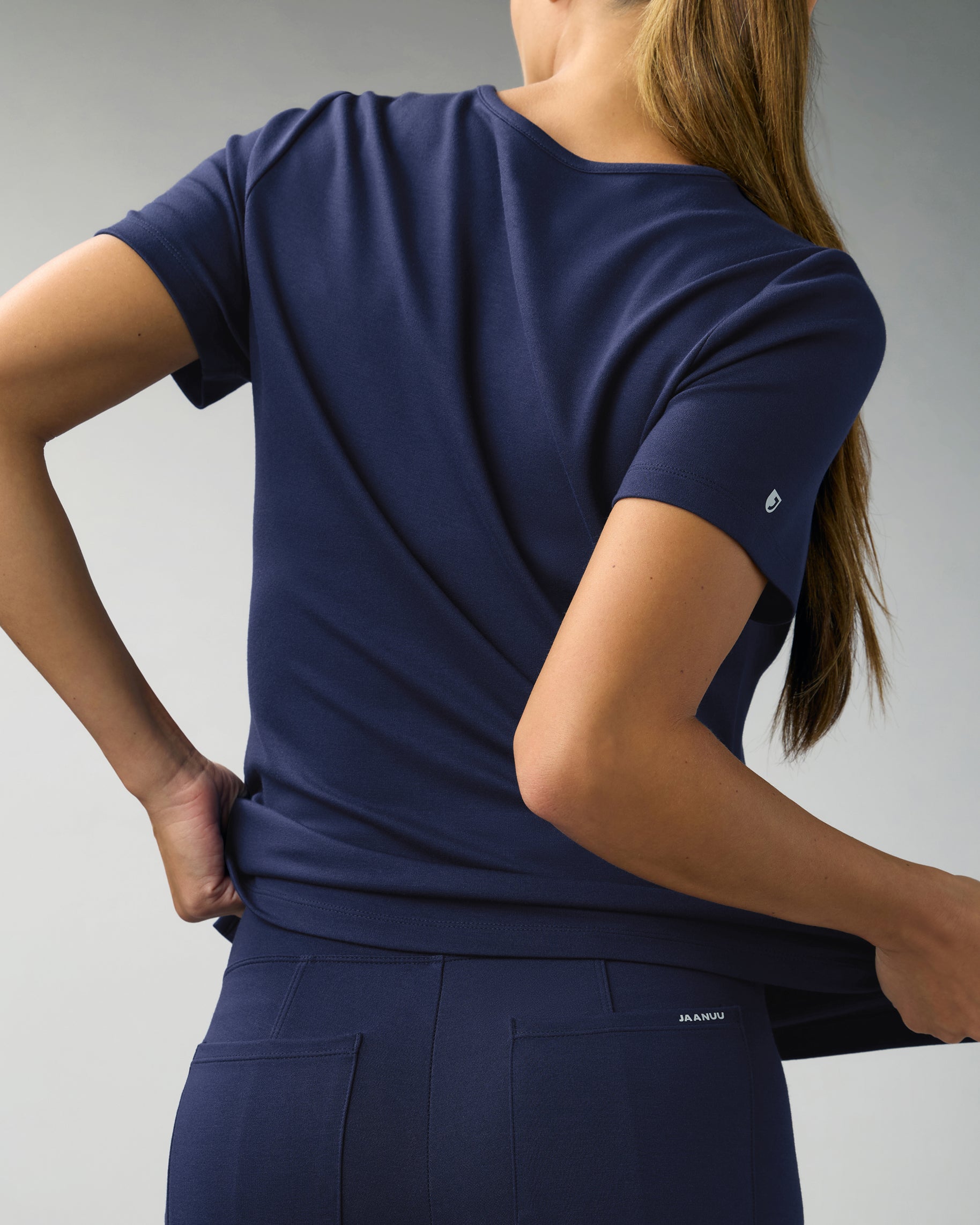 Back view of a woman stretching the hem of a navy scrub top, worn with matching scrub pants with back pockets