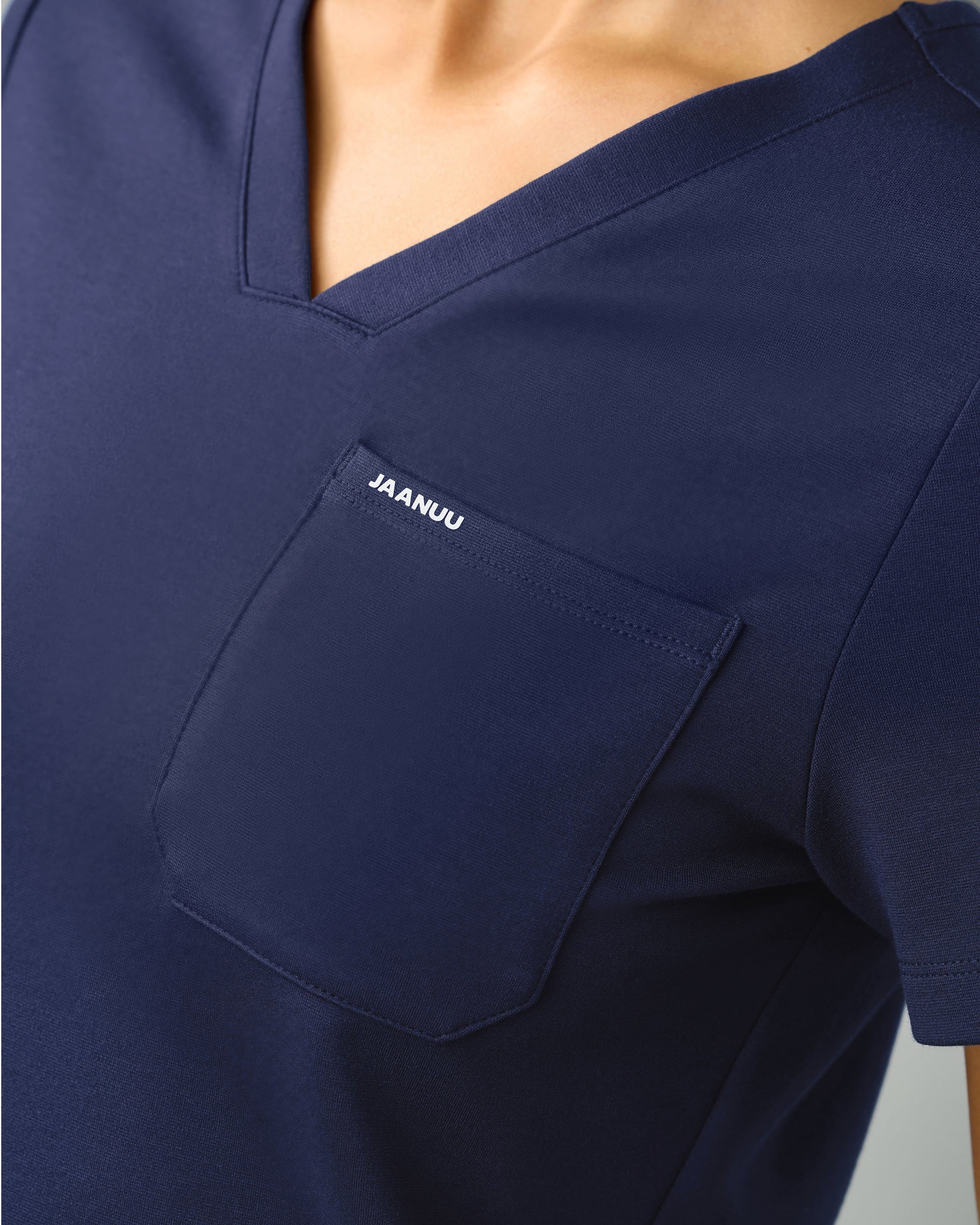 Close-up of the V-neckline and chest pocket marked JAANUU on a navy scrub top