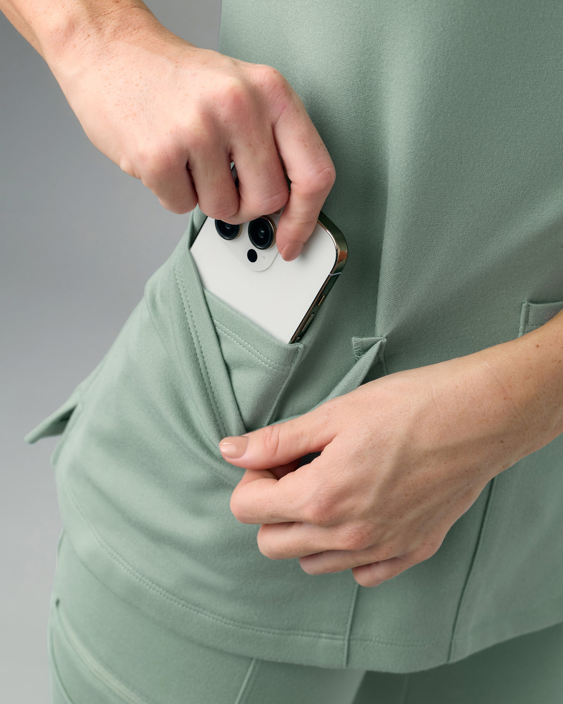 Close-up of a hand slipping a phone into the side patch pocket of a sage green scrub top