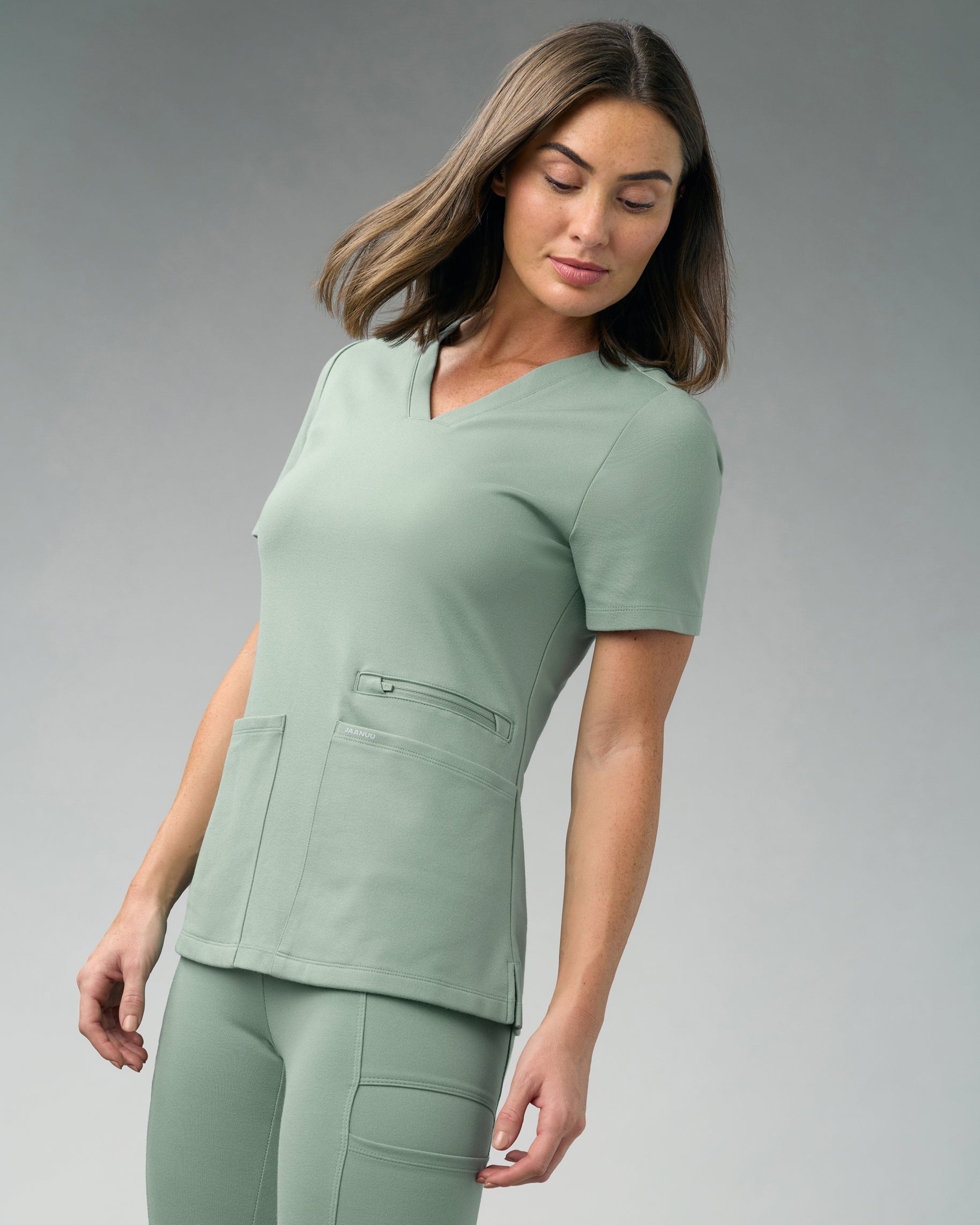 Woman in a sage green slim V-neck scrub top with a zippered pocket and two patch pockets, with matching scrub pants
