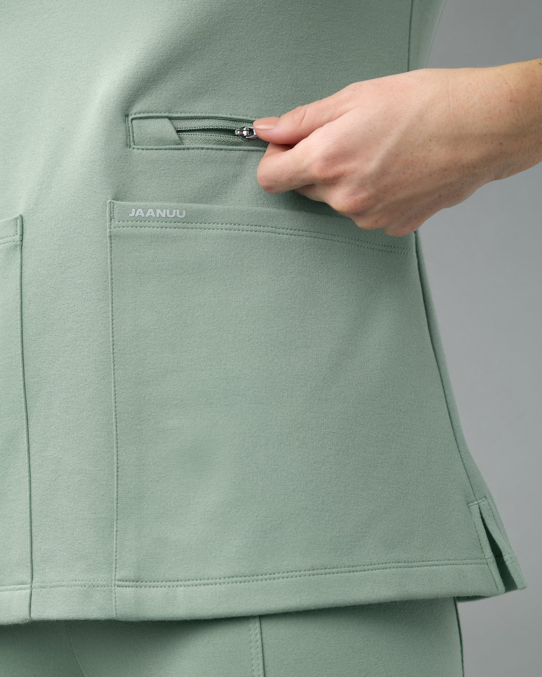Close-up of a hand opening the zippered pocket above a patch pocket marked JAANUU on a sage green scrub top