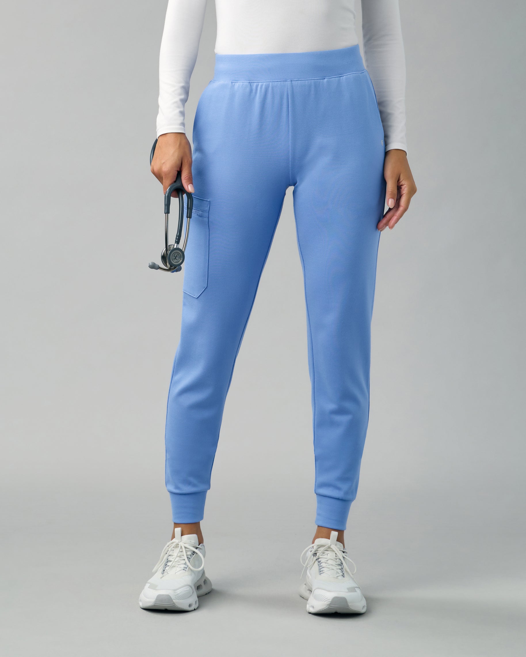 Front view of light blue slim scrub joggers with a side cargo pocket, worn with a white long-sleeve top and a stethoscope in one hand