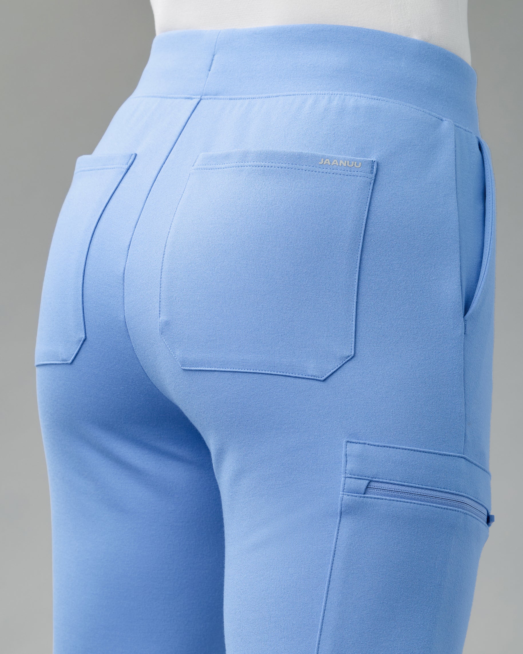 Back view of light blue scrub joggers showing two back patch pockets, a JAANUU label and a zippered cargo pocket
