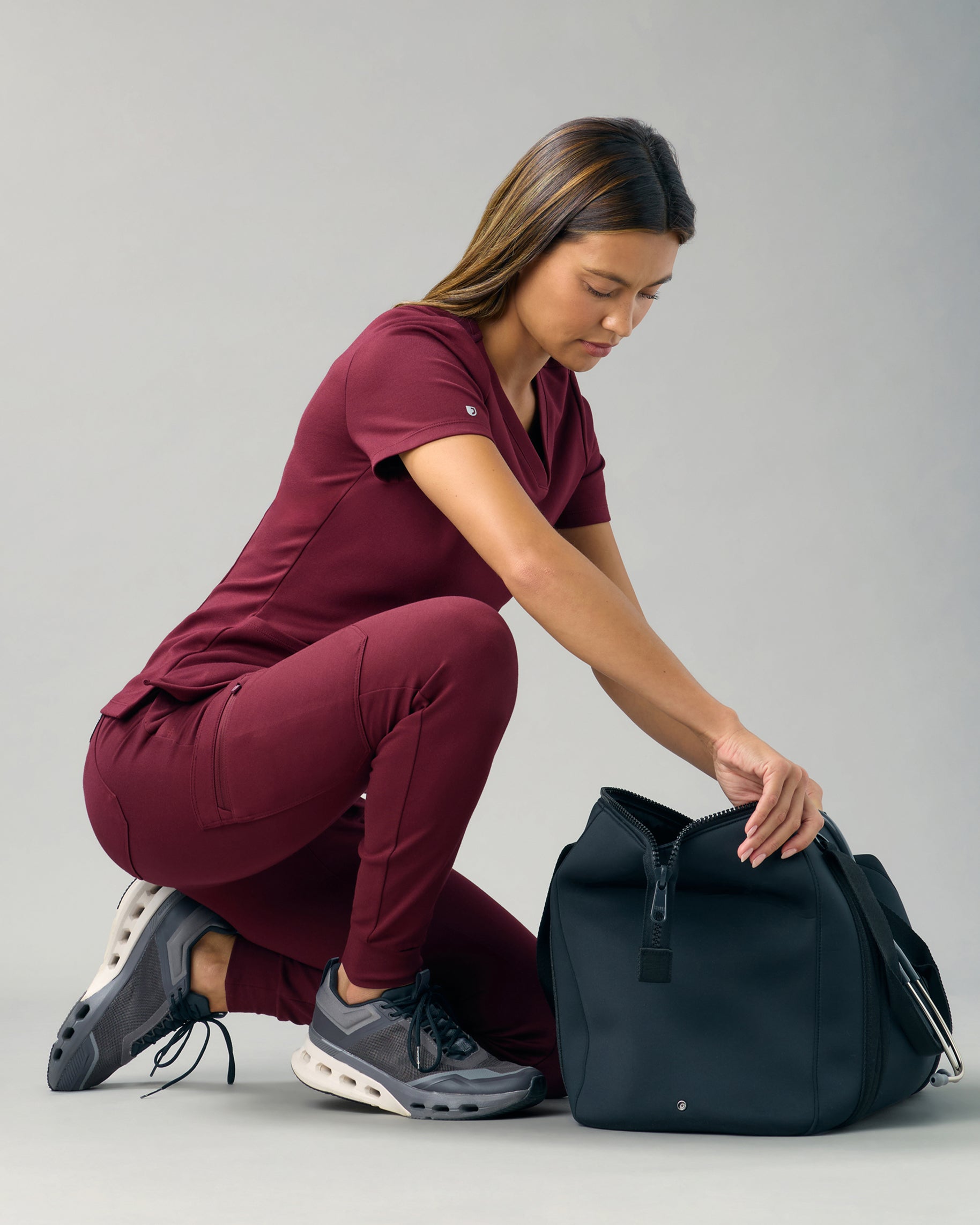Woman in a burgundy scrub top and skinny scrub joggers kneeling to open a black duffel bag