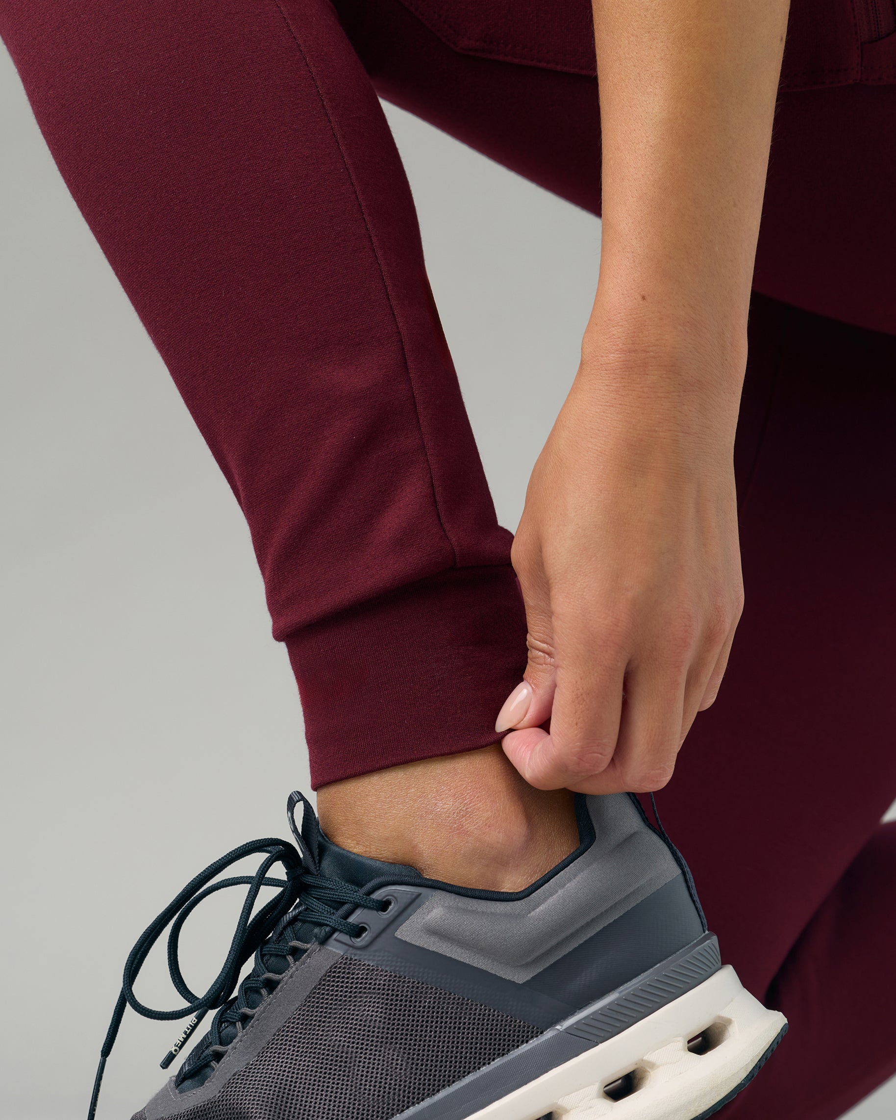 Close-up of a hand at the rib-knit ankle cuff of burgundy scrub joggers above a gray sneaker