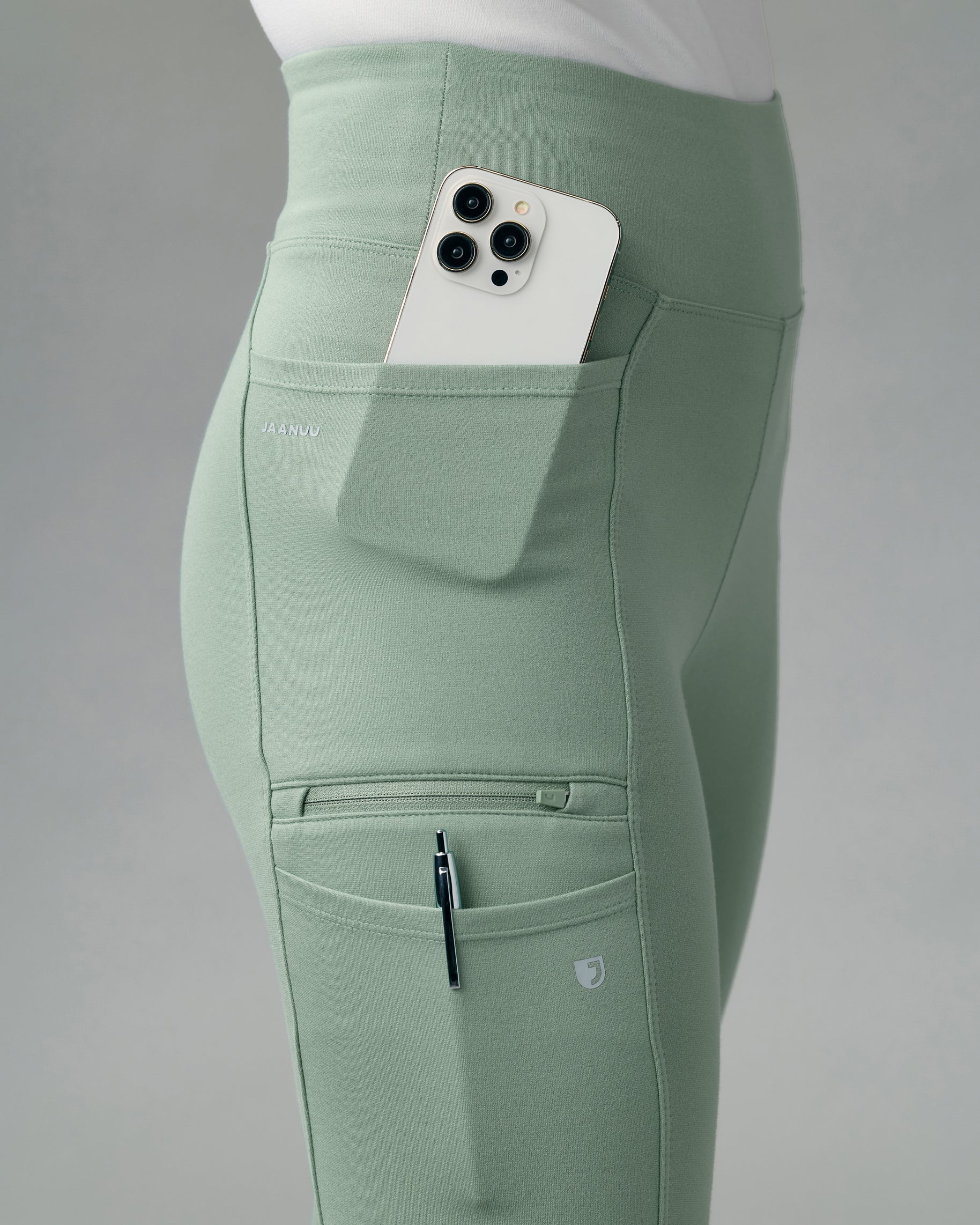 Close-up of the side pockets of sage green yoga scrub pants holding a phone and a pen, with a zippered pocket and JAANUU on the upper pocket