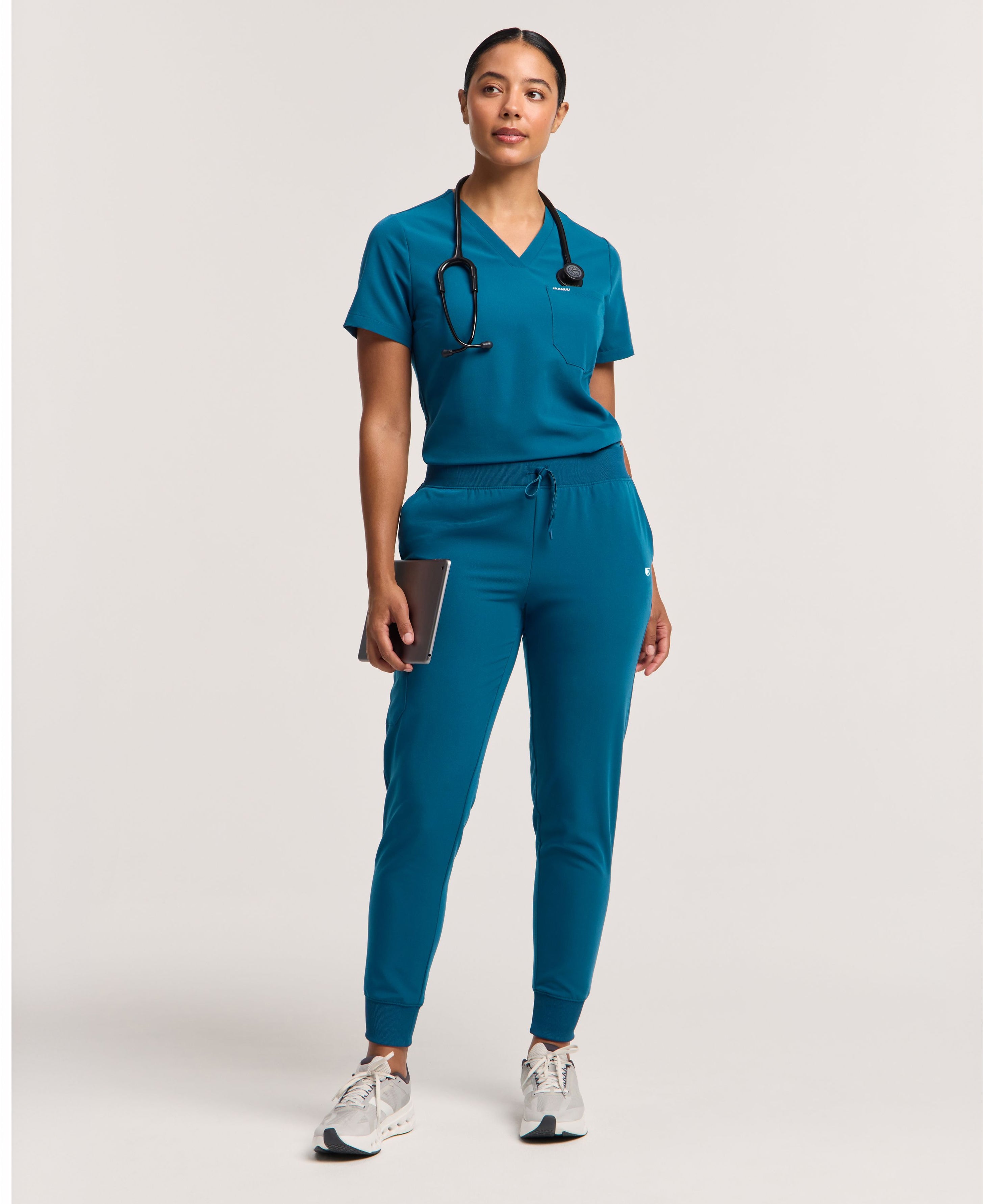 Woman in a Caribbean Blue V-neck scrub top with a chest pocket and matching scrub joggers, with a stethoscope around her neck and a tablet in one hand