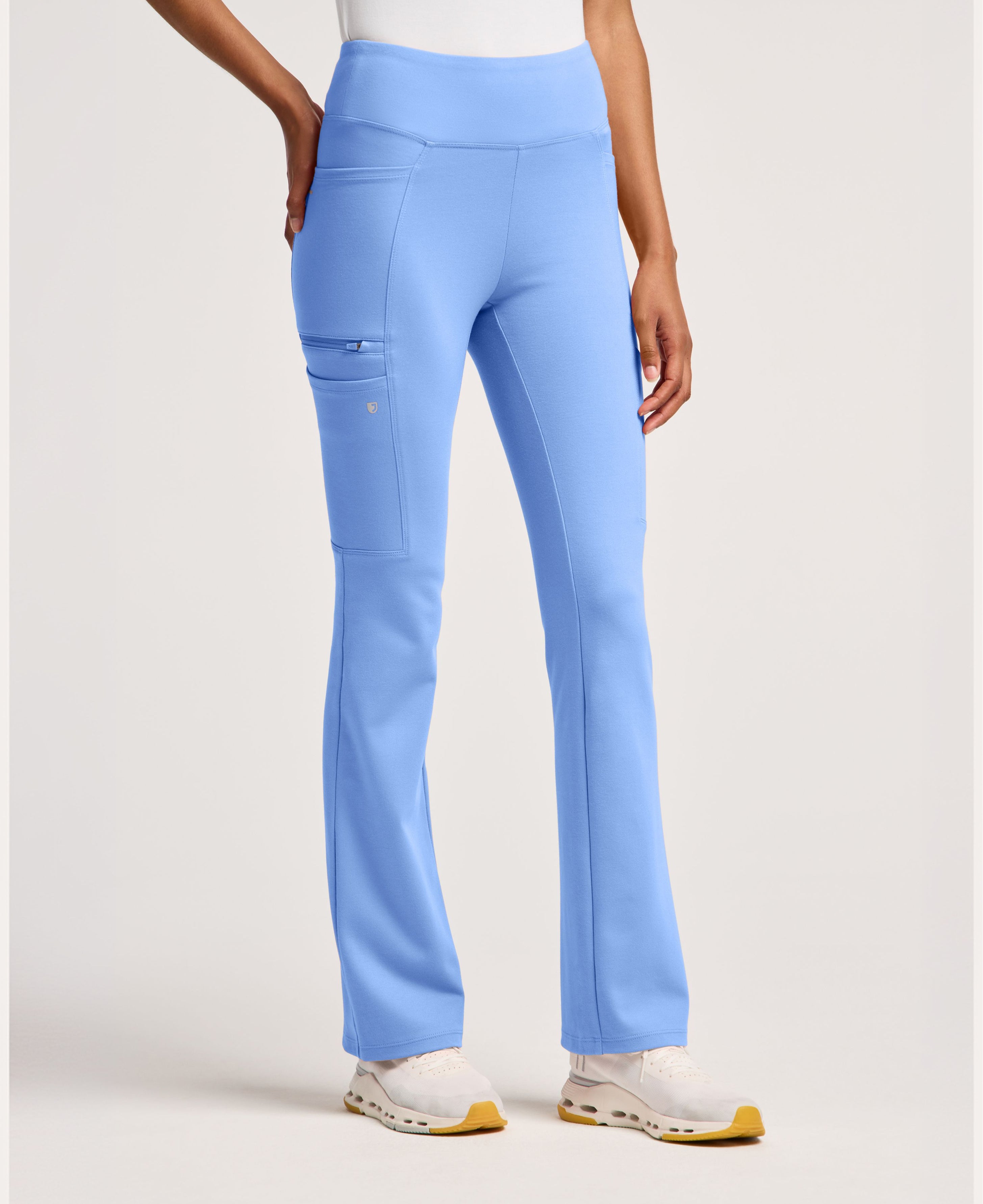 Front view of Ceil Blue yoga scrub pants with a high waistband, a zippered side cargo pocket and a flared leg, worn with a white top and white sneakers