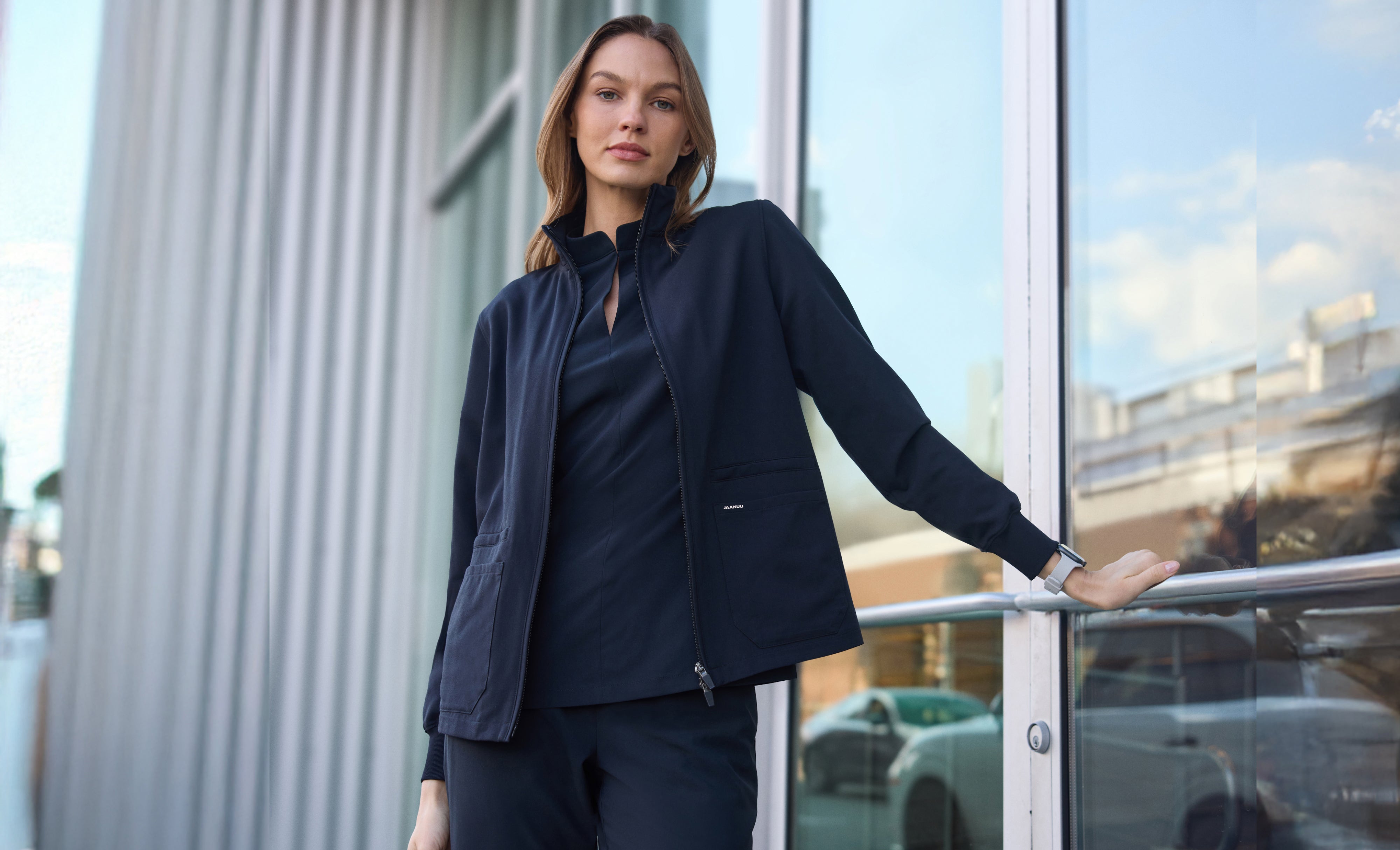 Woman in a black Jaanuu zip-up scrub jacket over a matching stand-collar scrub top and scrub pants, resting one hand on a railing outside a glass building entrance