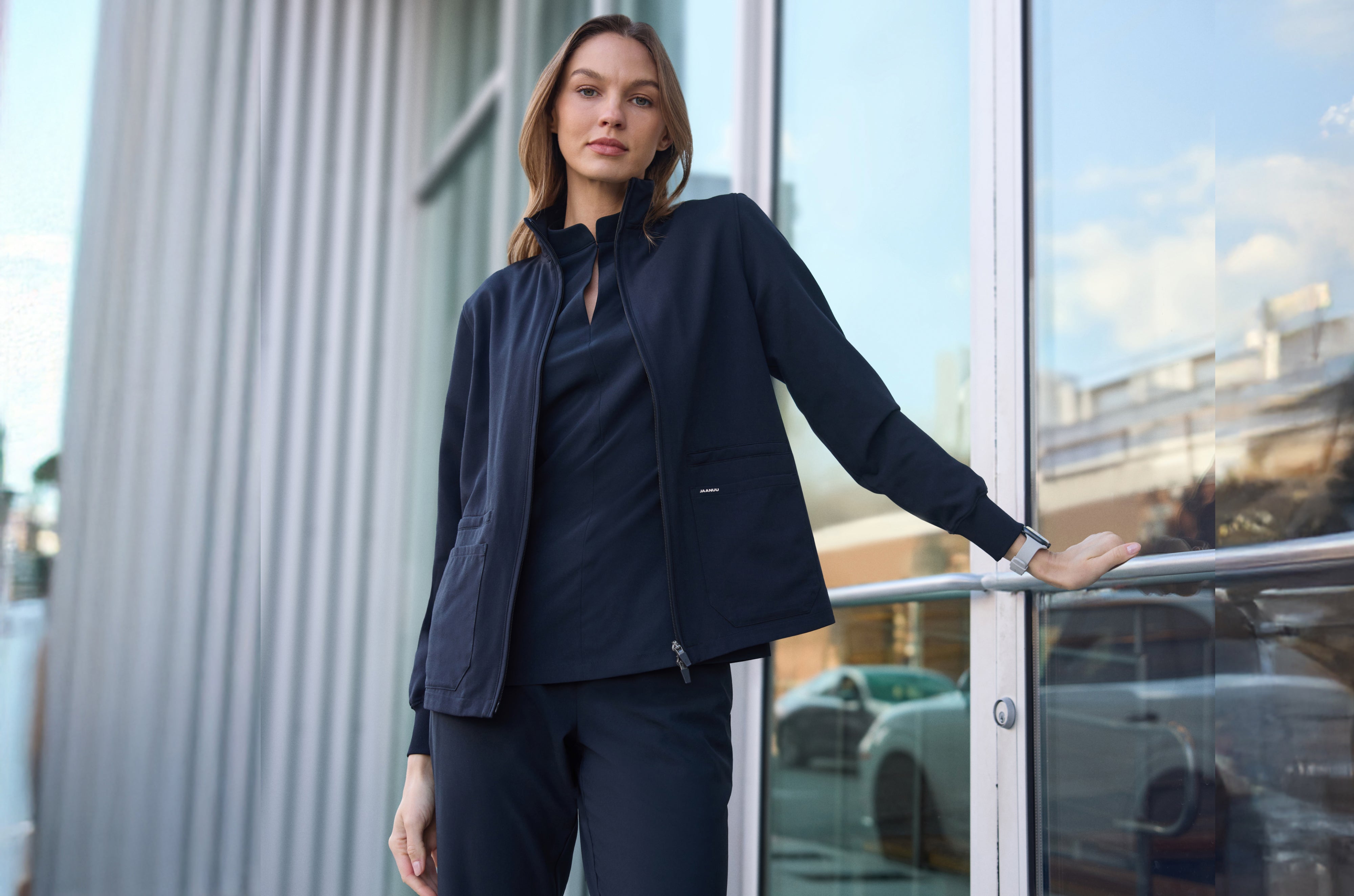 Woman in a black Jaanuu zip-up scrub jacket over a matching stand-collar scrub top and scrub pants, resting one hand on a railing outside a glass building entrance