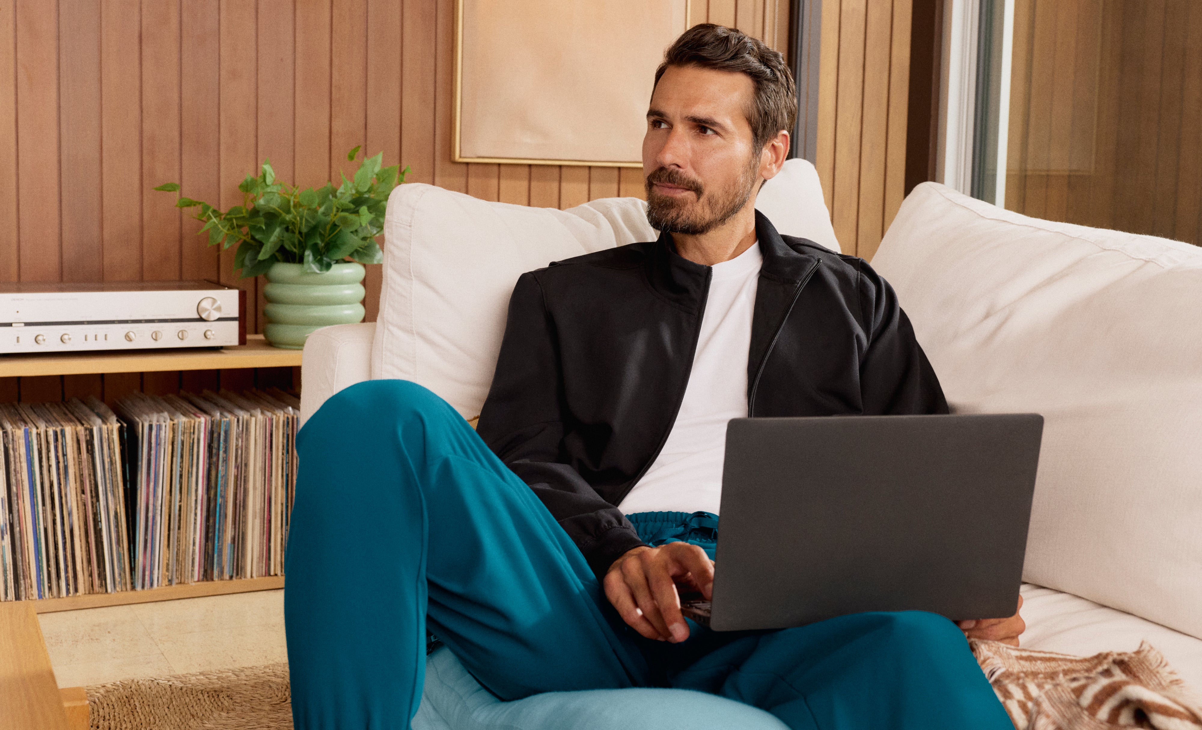 Man in a black Jaanuu zip-up jacket over a white tee and teal jogger scrub pants, sitting on a white sofa with a laptop beside a shelf of vinyl records