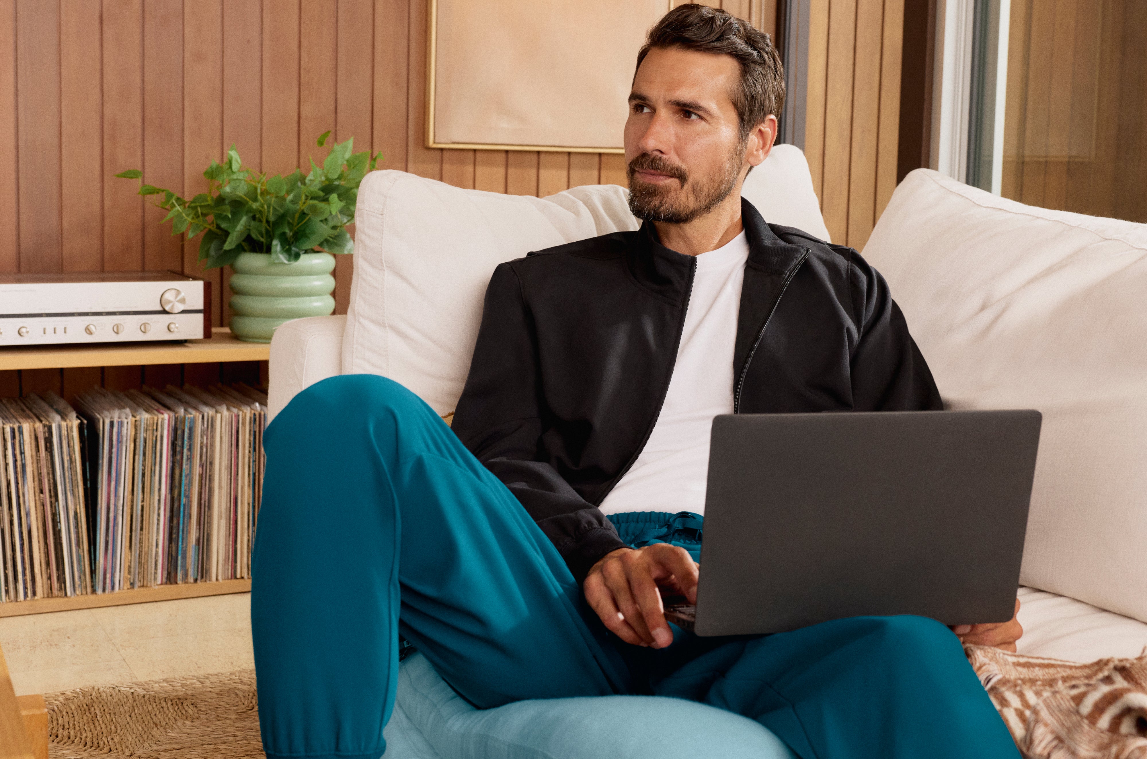 Man in a black Jaanuu zip-up jacket over a white tee and teal jogger scrub pants, sitting on a white sofa with a laptop beside a shelf of vinyl records