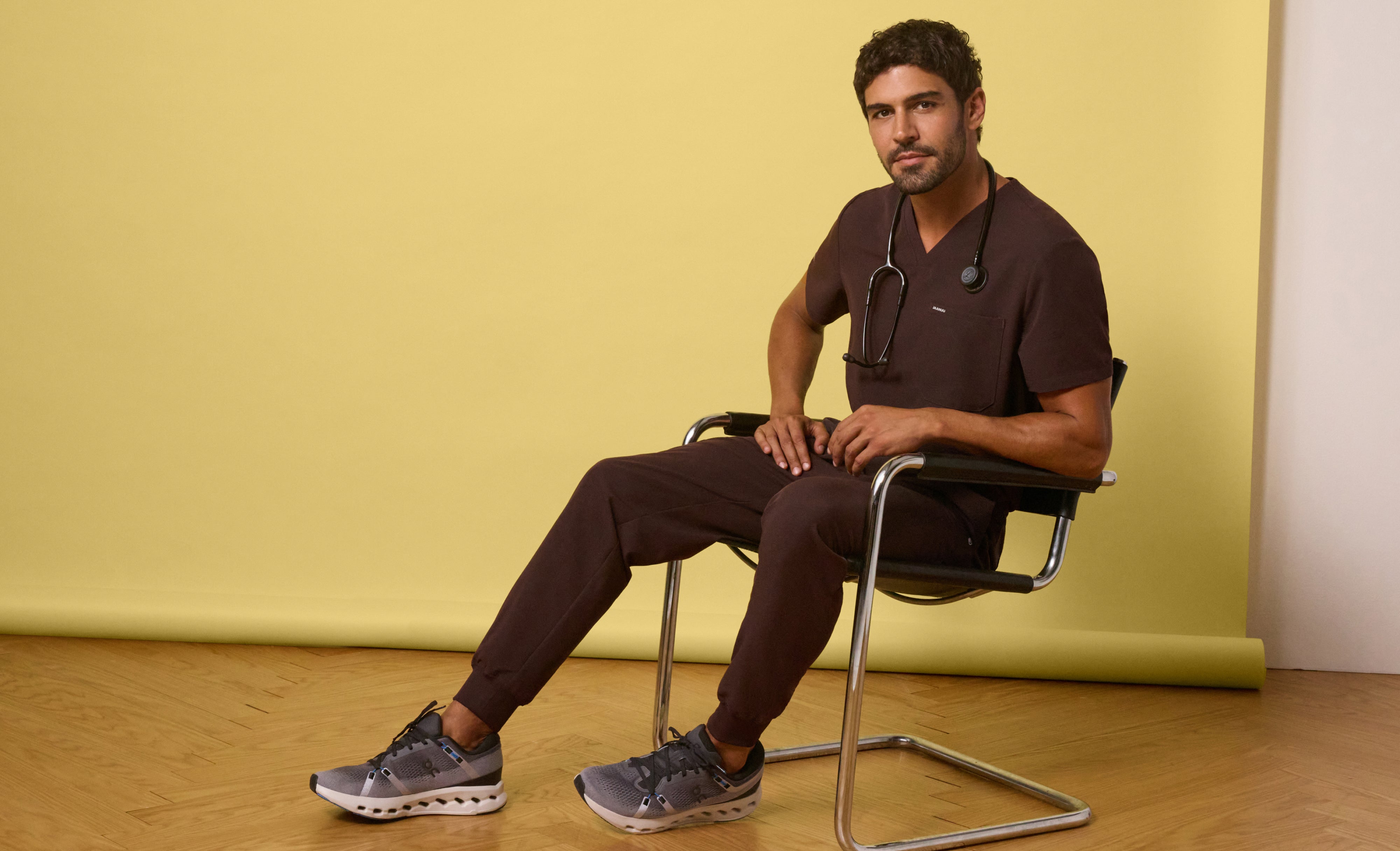 Man in an Espresso (dark brown) Jaanuu V-neck scrub top and jogger scrub pants with a stethoscope around his neck, sitting in a chrome chair against a yellow backdrop