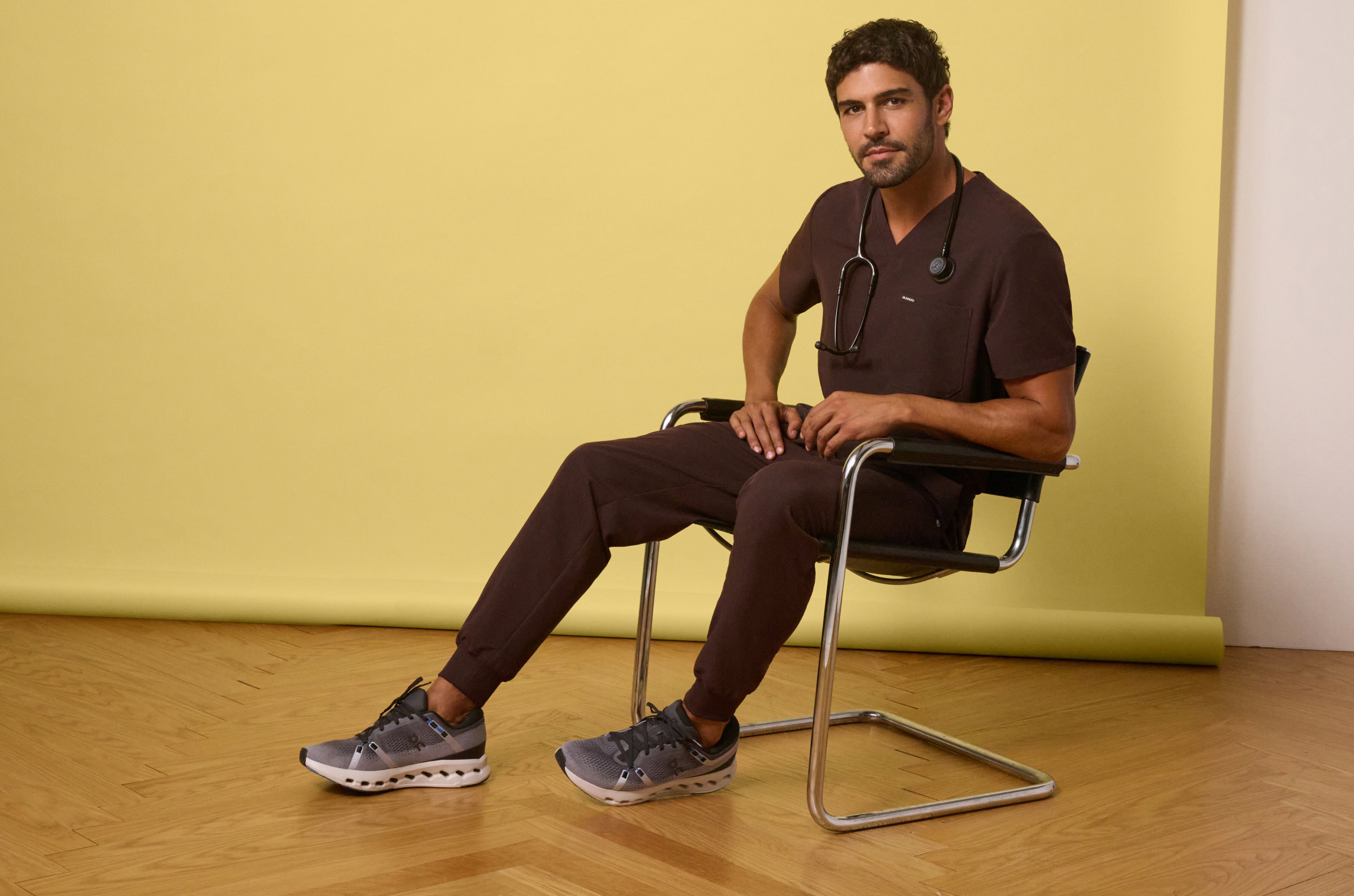Man in an Espresso (dark brown) Jaanuu V-neck scrub top and jogger scrub pants with a stethoscope around his neck, sitting in a chrome chair against a yellow backdrop