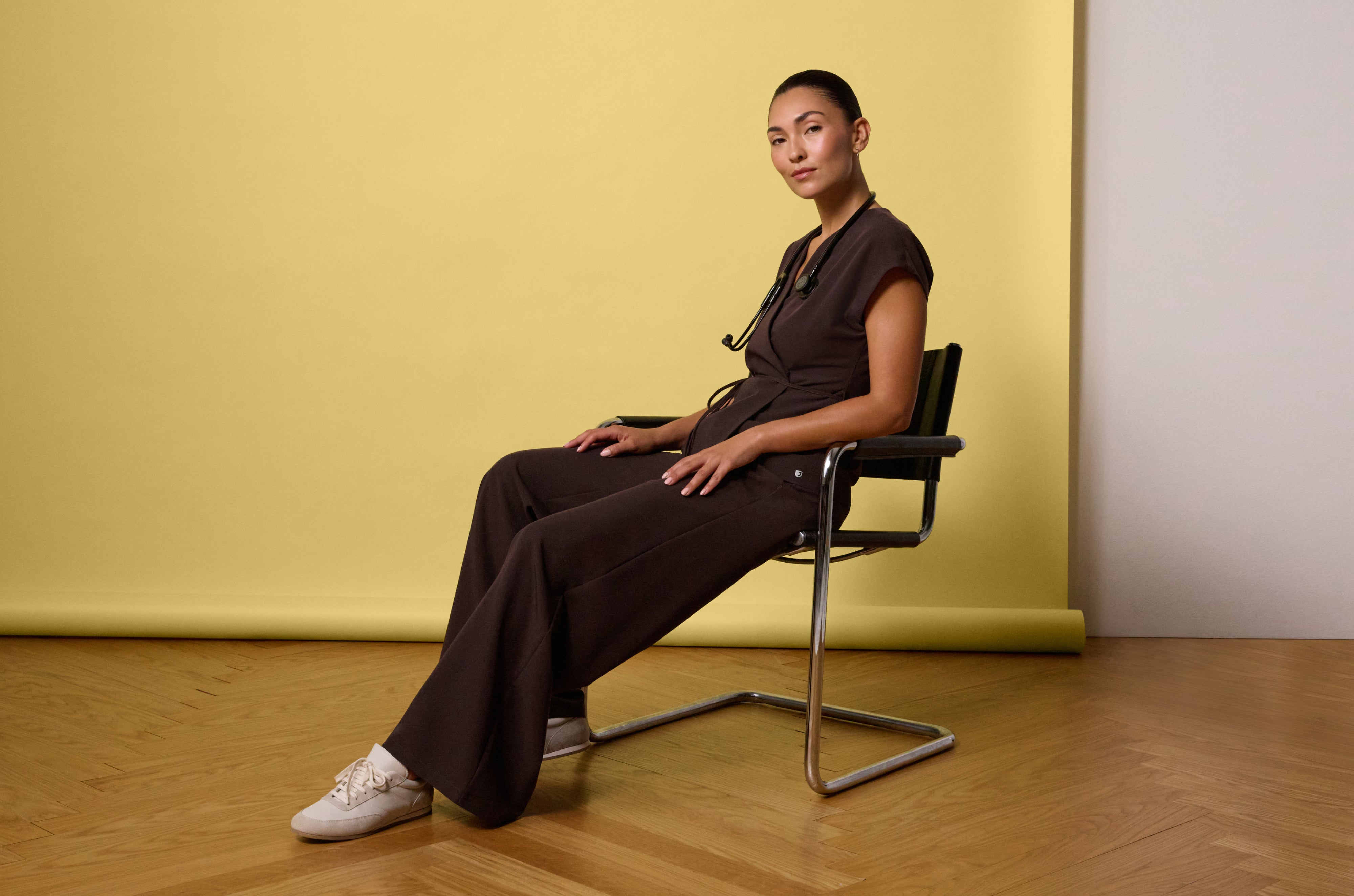 Woman in an Espresso (dark brown) Jaanuu wrap-front scrub top with a tie waist and wide-leg scrub pants, with a stethoscope around her neck, sitting in a chrome chair against a yellow backdrop