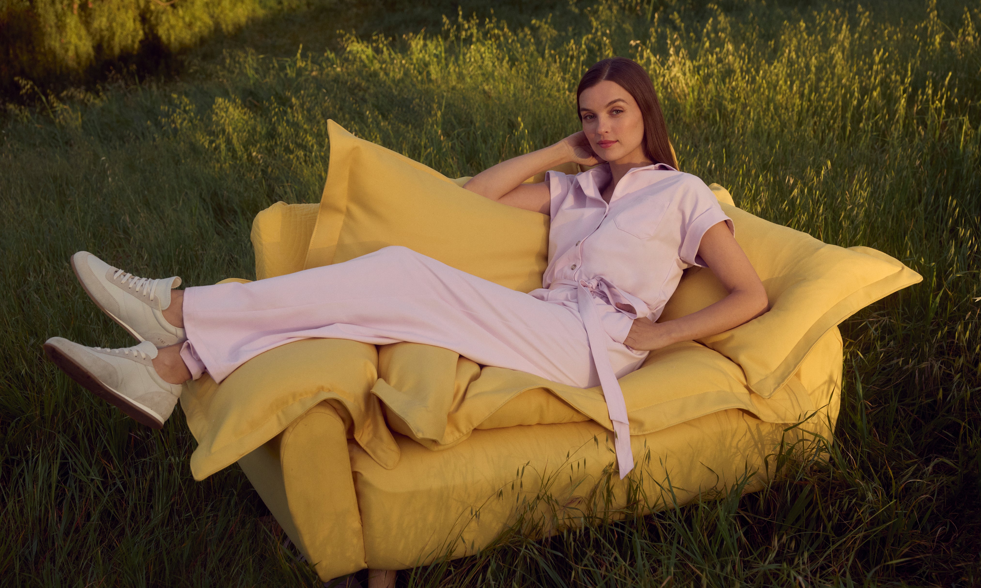 Woman in a pale pink Jaanuu short-sleeve belted jumpsuit reclining on a yellow armchair set in a grassy field at golden hour