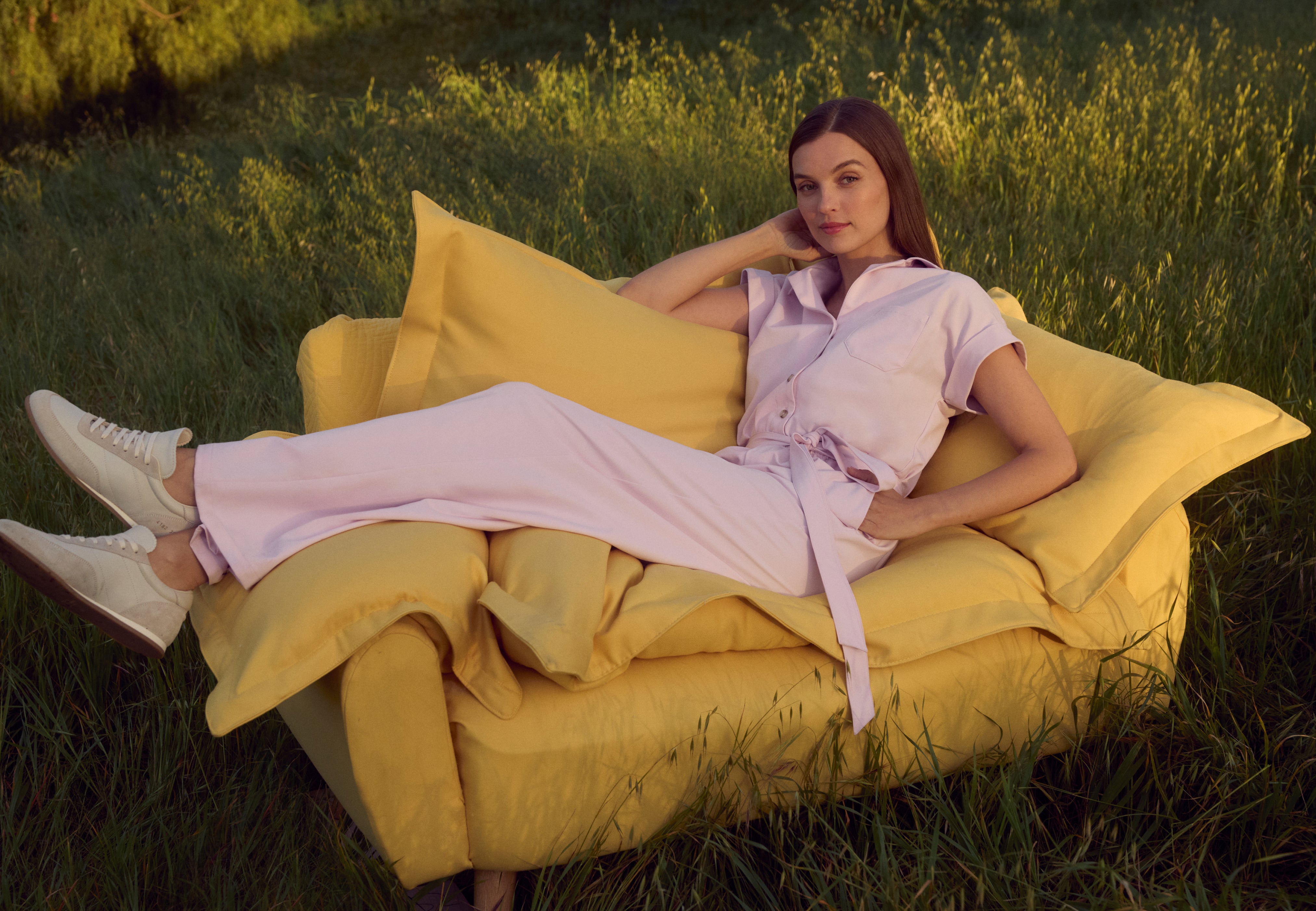 Woman in a pale pink Jaanuu short-sleeve belted jumpsuit reclining on a yellow armchair set in a grassy field at golden hour