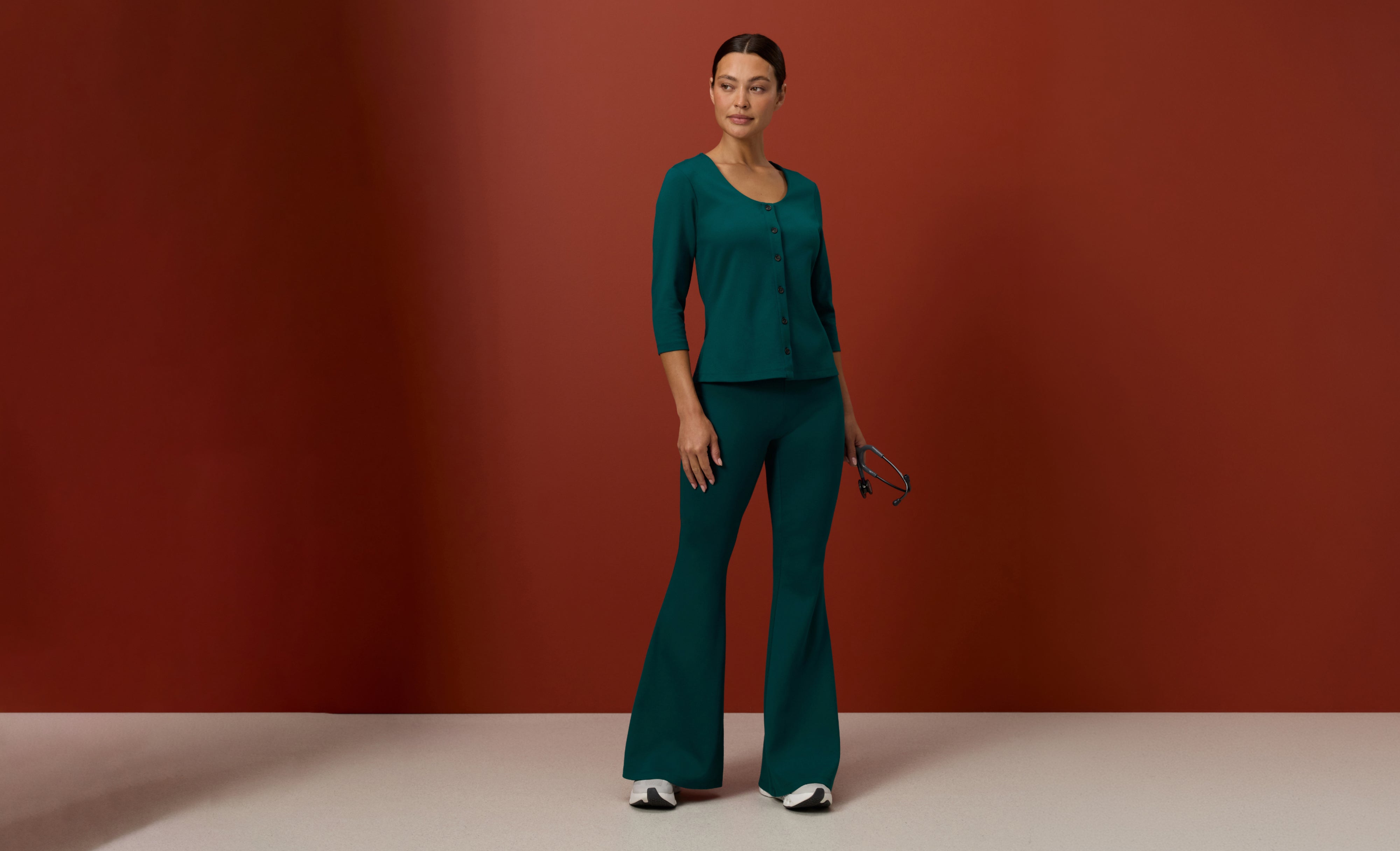 Woman in a Midnight Green (deep teal green) Jaanuu button-front scrub top with three-quarter sleeves and flared scrub pants, holding a stethoscope against a rust red wall