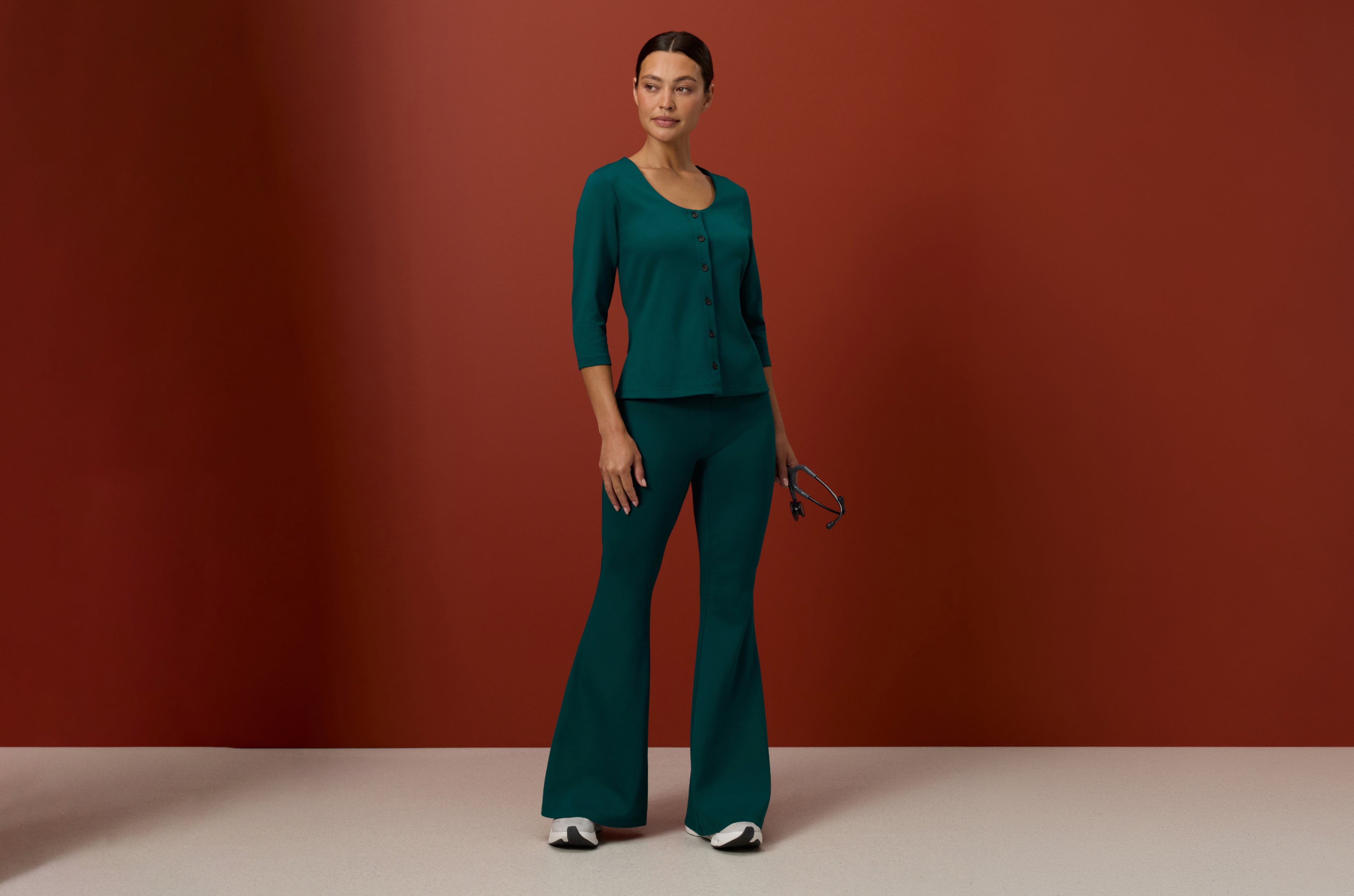 Woman in a Midnight Green (deep teal green) Jaanuu button-front scrub top with three-quarter sleeves and flared scrub pants, holding a stethoscope against a rust red wall