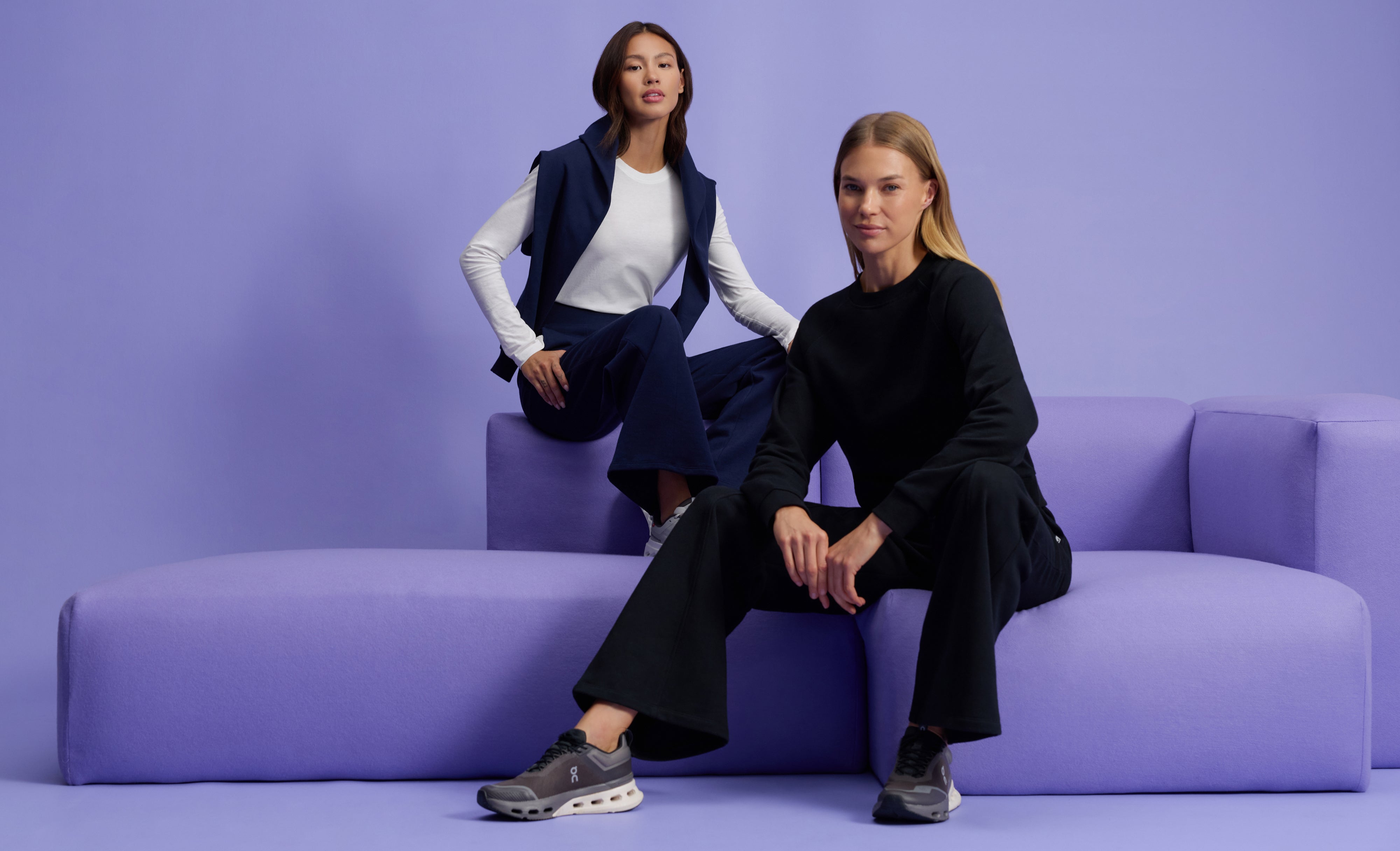 Two women in Jaanuu loungewear on a lavender sofa, one in a white long-sleeve tee and navy wide-leg pants with a navy sweatshirt over her shoulders, the other in a black crewneck sweatshirt and black wide-leg pants