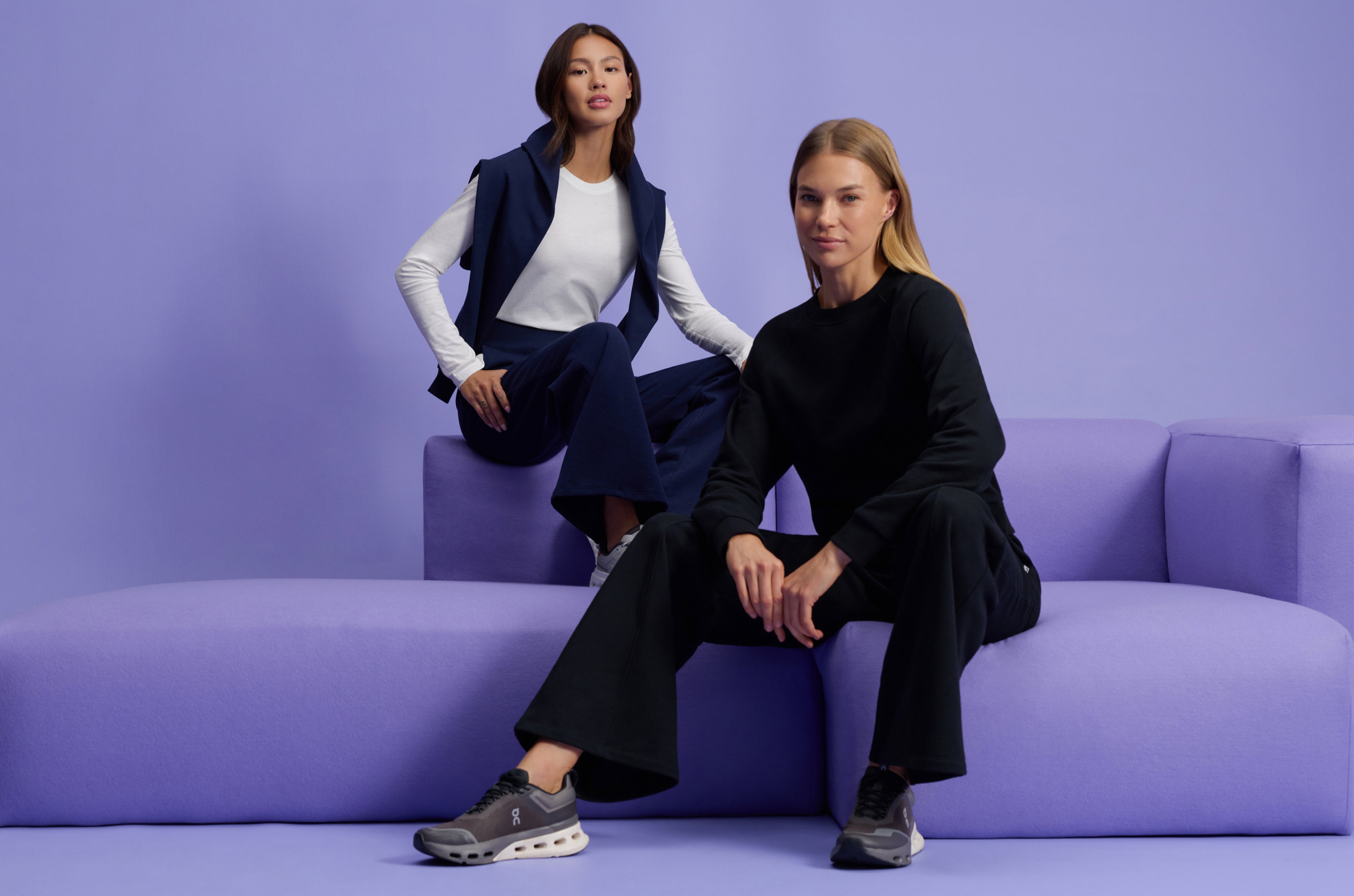 Two women in Jaanuu loungewear on a lavender sofa, one in a white long-sleeve tee and navy wide-leg pants with a navy sweatshirt over her shoulders, the other in a black crewneck sweatshirt and black wide-leg pants