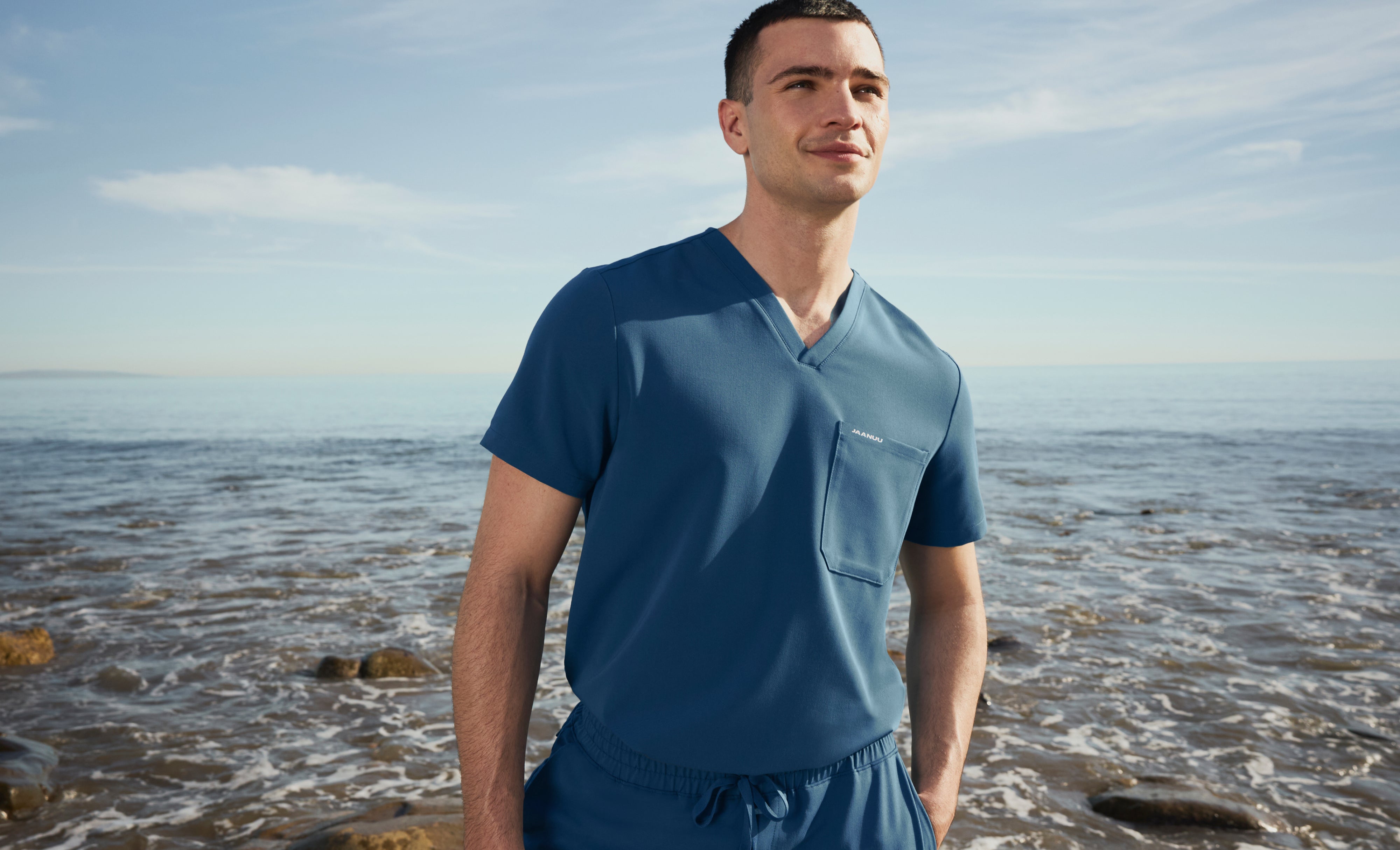 Man in an Ocean (medium blue) Jaanuu V-neck scrub top and drawstring scrub pants with one hand in his pocket, standing on a rocky shore with waves behind him
