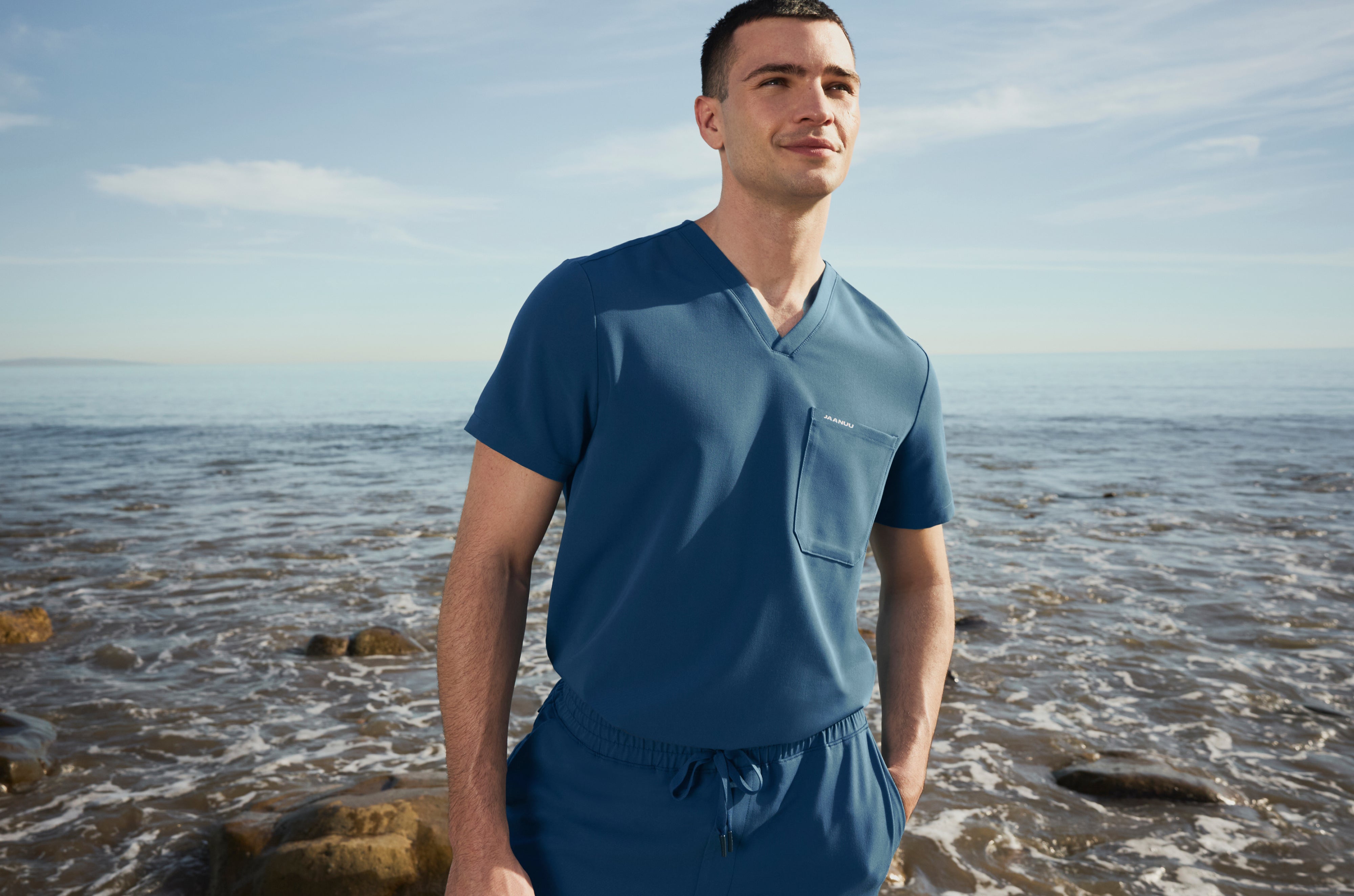 Man in an Ocean (medium blue) Jaanuu V-neck scrub top and drawstring scrub pants with one hand in his pocket, standing on a rocky shore with waves behind him