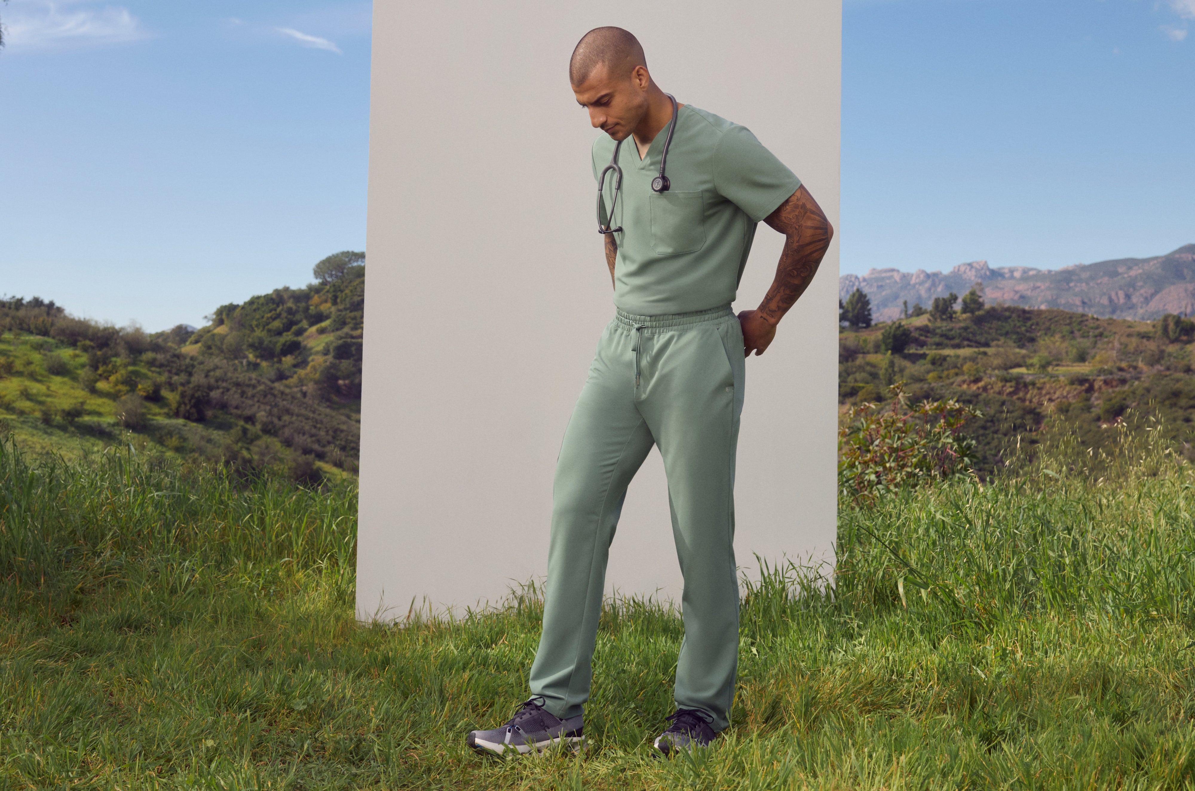 Man in a Sage (soft green) Jaanuu V-neck scrub top and scrub pants with a stethoscope around his neck, looking down as he stands in tall grass in front of a white panel with green hills behind him