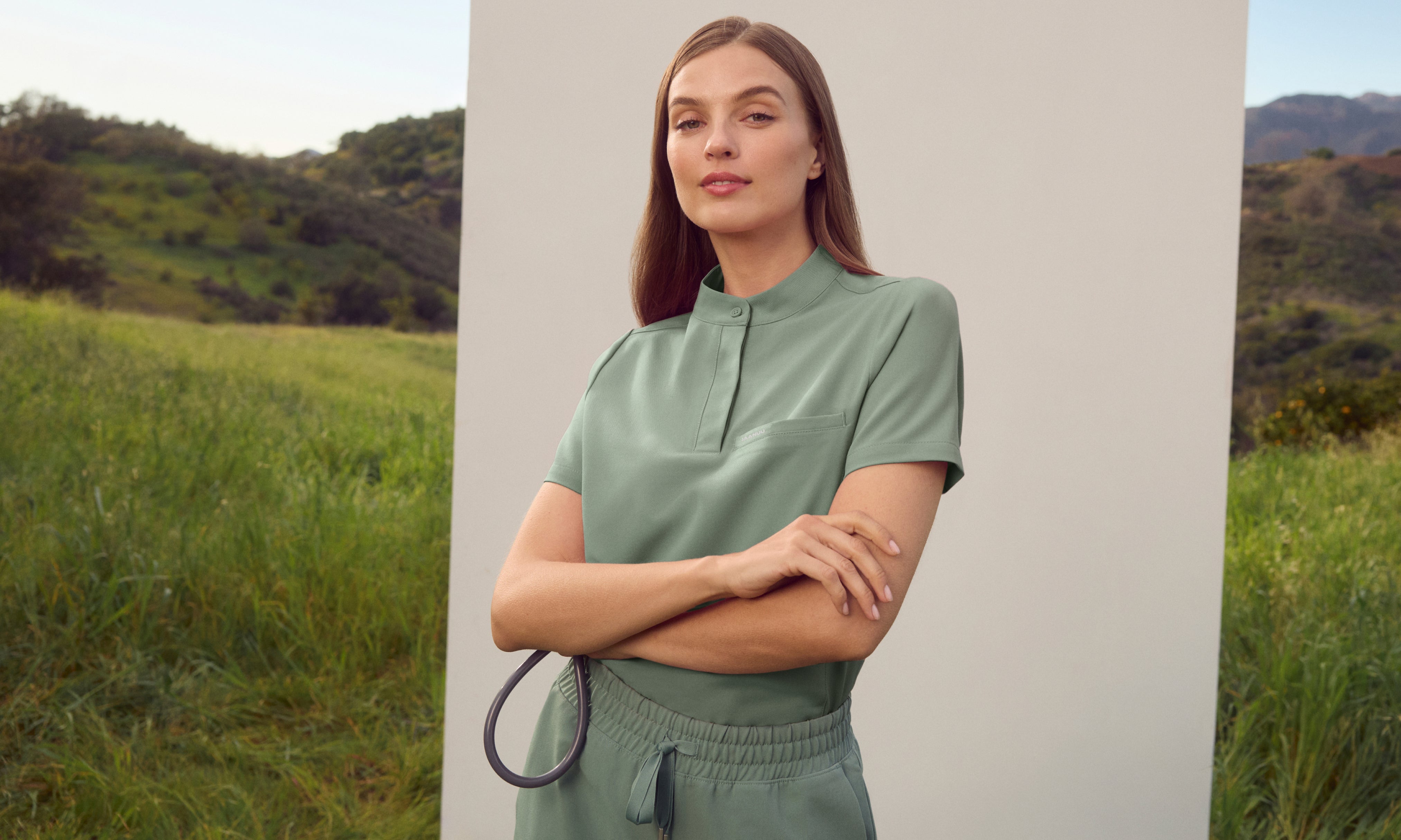 Woman in a Sage (soft green) Jaanuu stand-collar scrub top and drawstring scrub pants with her arms crossed, holding a stethoscope in front of a white panel in a grassy field with green hills behind her