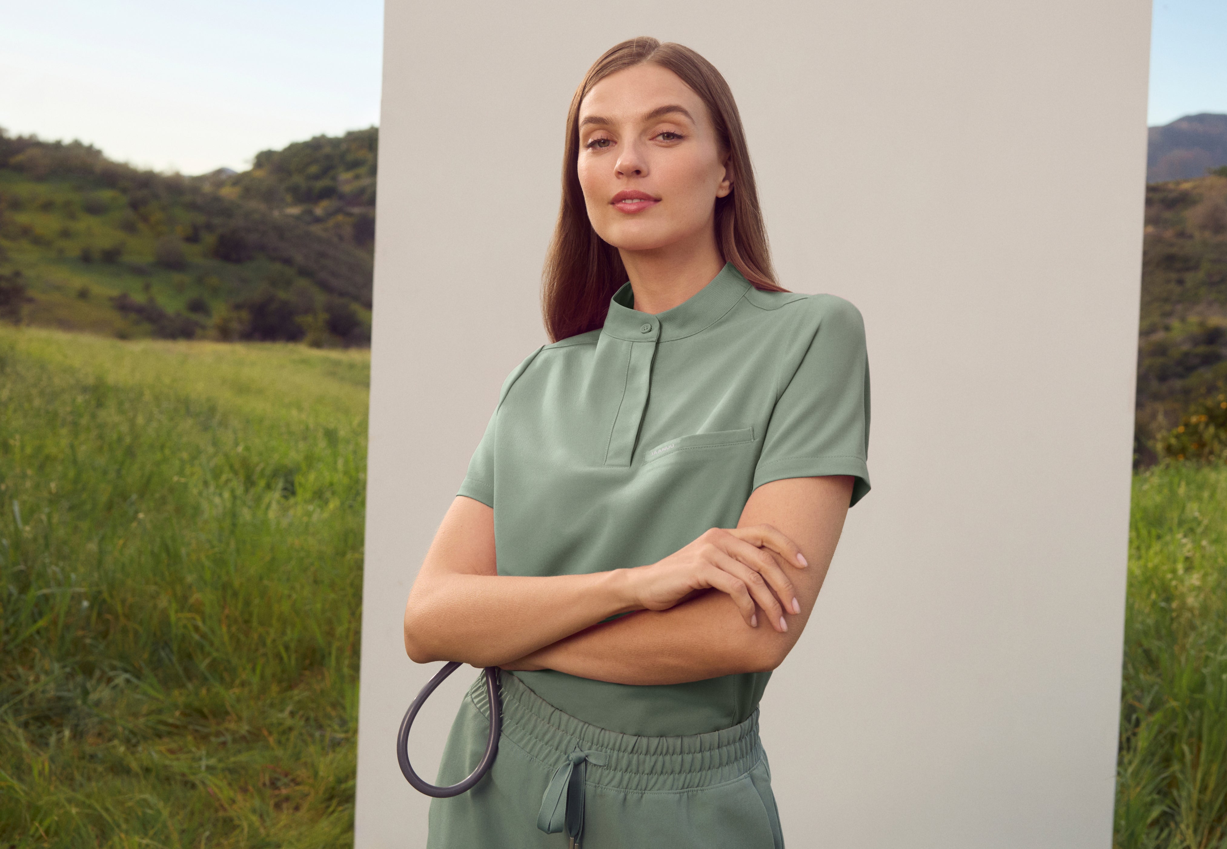 Woman in a Sage (soft green) Jaanuu stand-collar scrub top and drawstring scrub pants with her arms crossed, holding a stethoscope in front of a white panel in a grassy field with green hills behind her