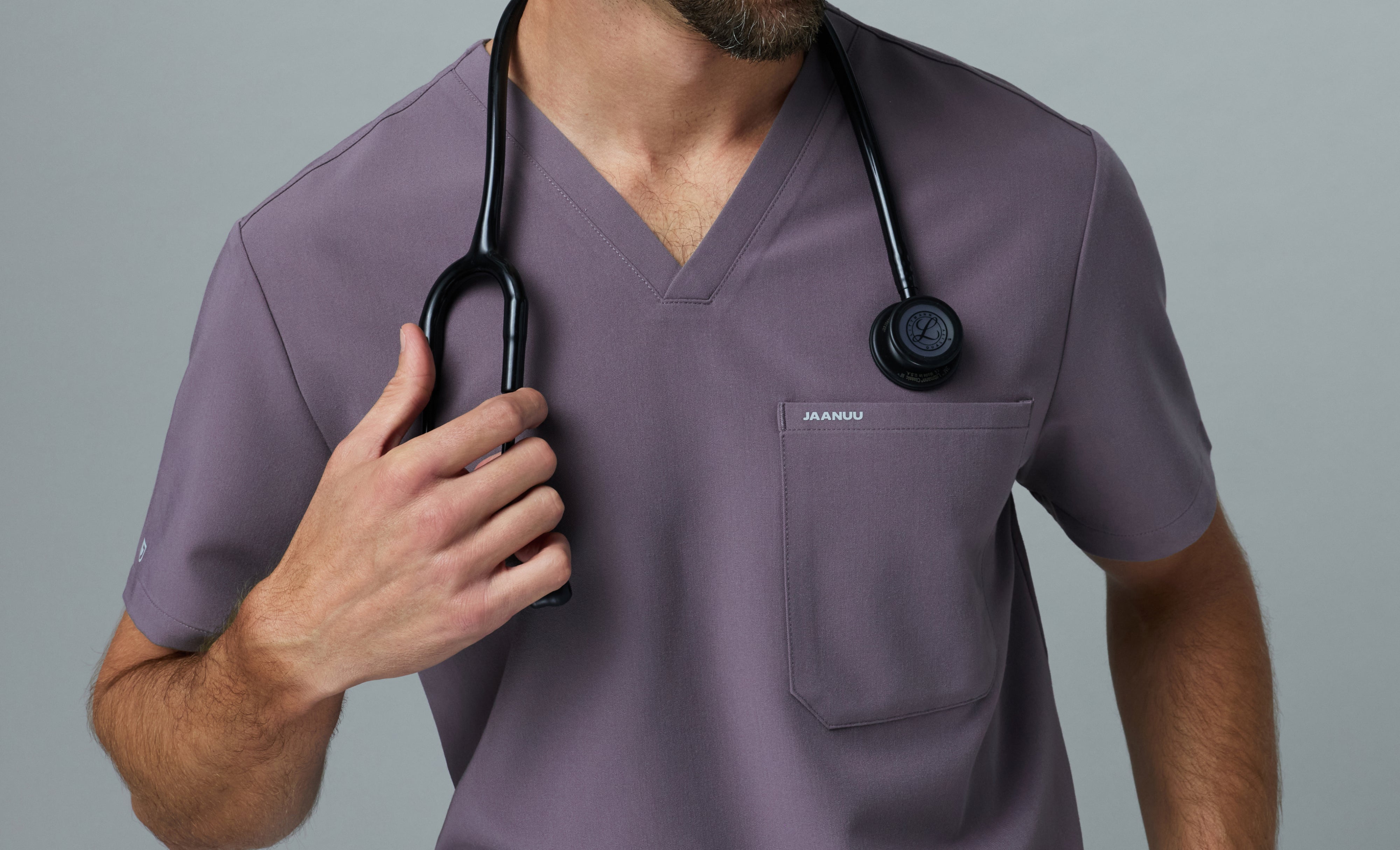 Close-up of a man in a Titanium (purple gray) Jaanuu V-neck scrub top with a chest pocket, holding the black stethoscope around his neck against a gray backdrop