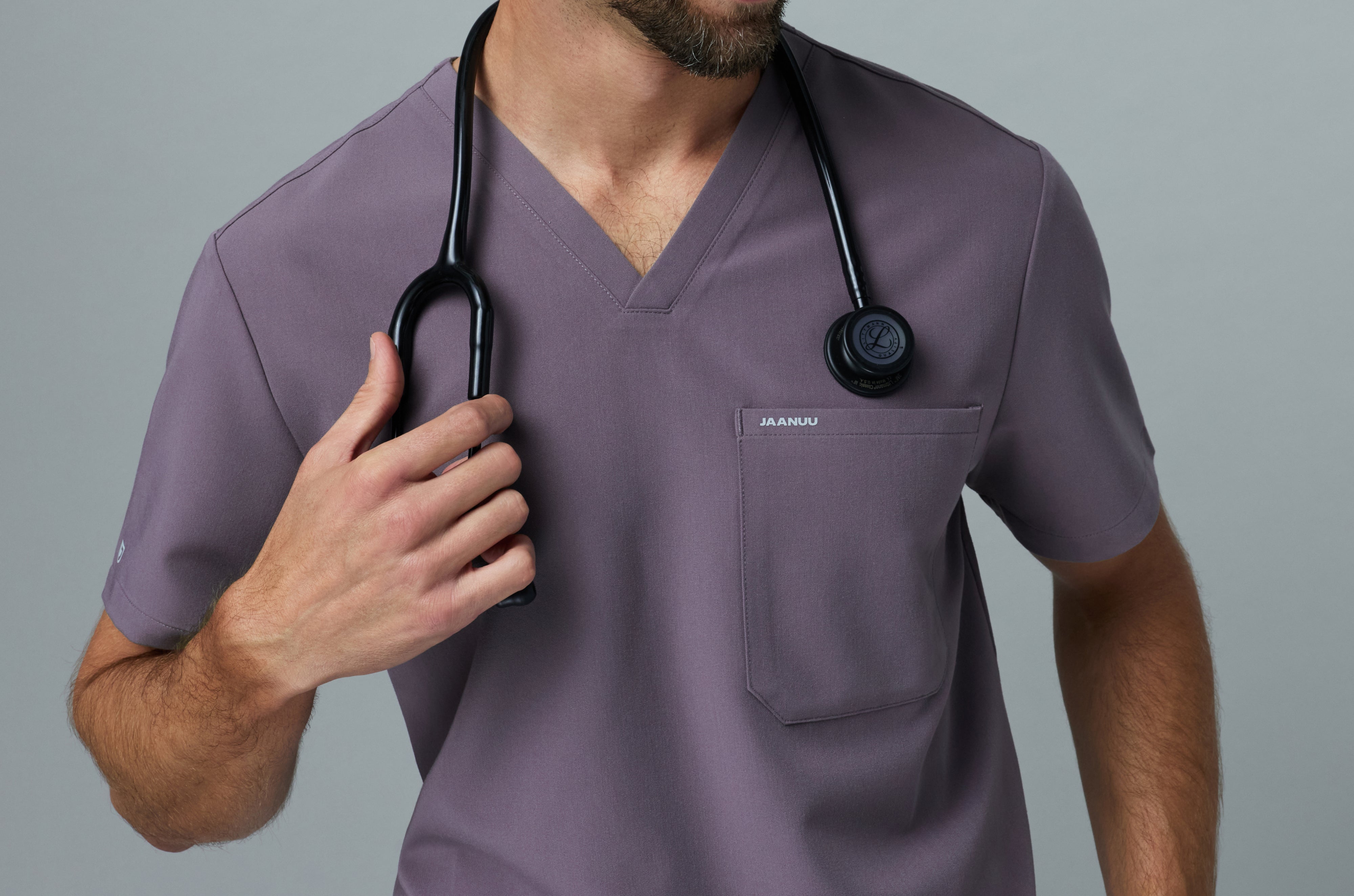 Close-up of a man in a Titanium (purple gray) Jaanuu V-neck scrub top with a chest pocket, holding the black stethoscope around his neck against a gray backdrop