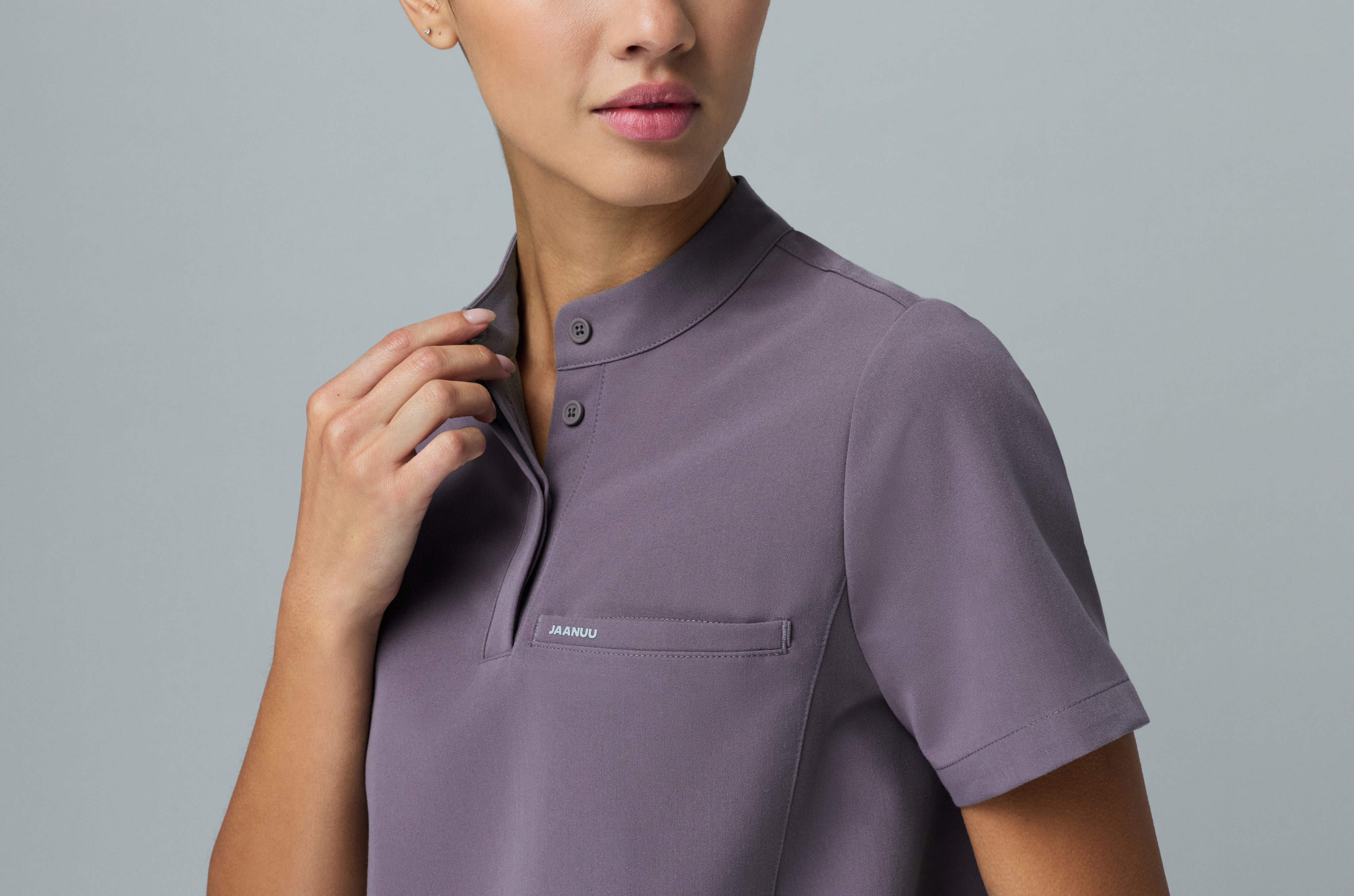 Close-up of a woman in a Titanium (purple gray) Jaanuu stand-collar scrub top with a buttoned placket and a chest pocket, touching the collar against a gray backdrop