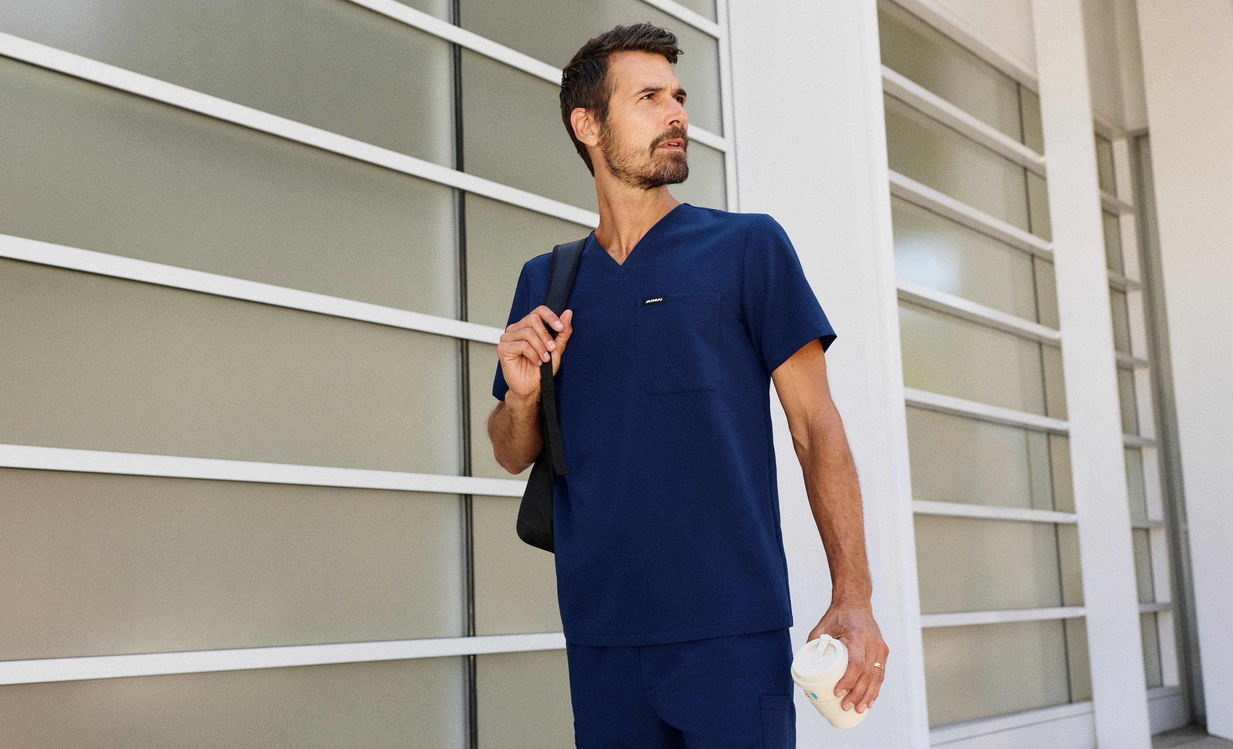 Man in a navy blue Jaanuu V-neck scrub top and matching scrub pants with a black bag over one shoulder and a coffee cup in hand, outside a building with frosted glass panels