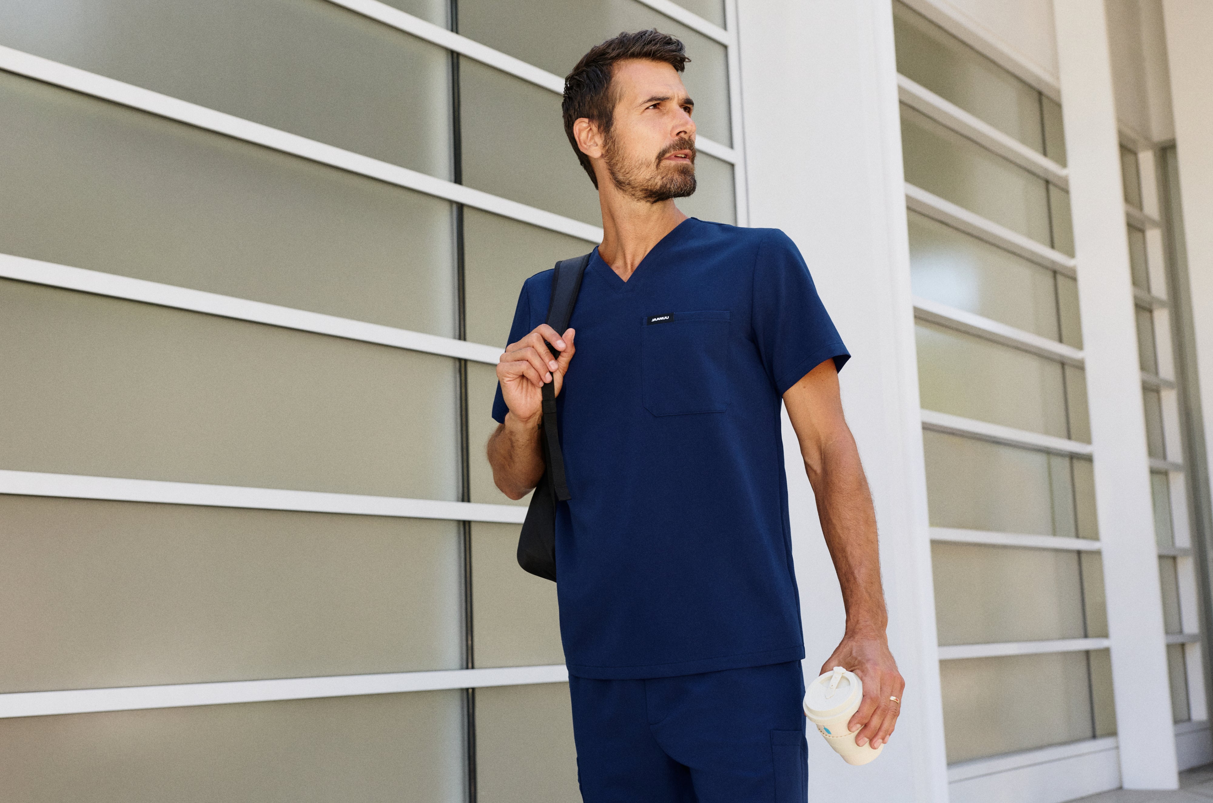 Man in a navy blue Jaanuu V-neck scrub top and matching scrub pants with a black bag over one shoulder and a coffee cup in hand, outside a building with frosted glass panels
