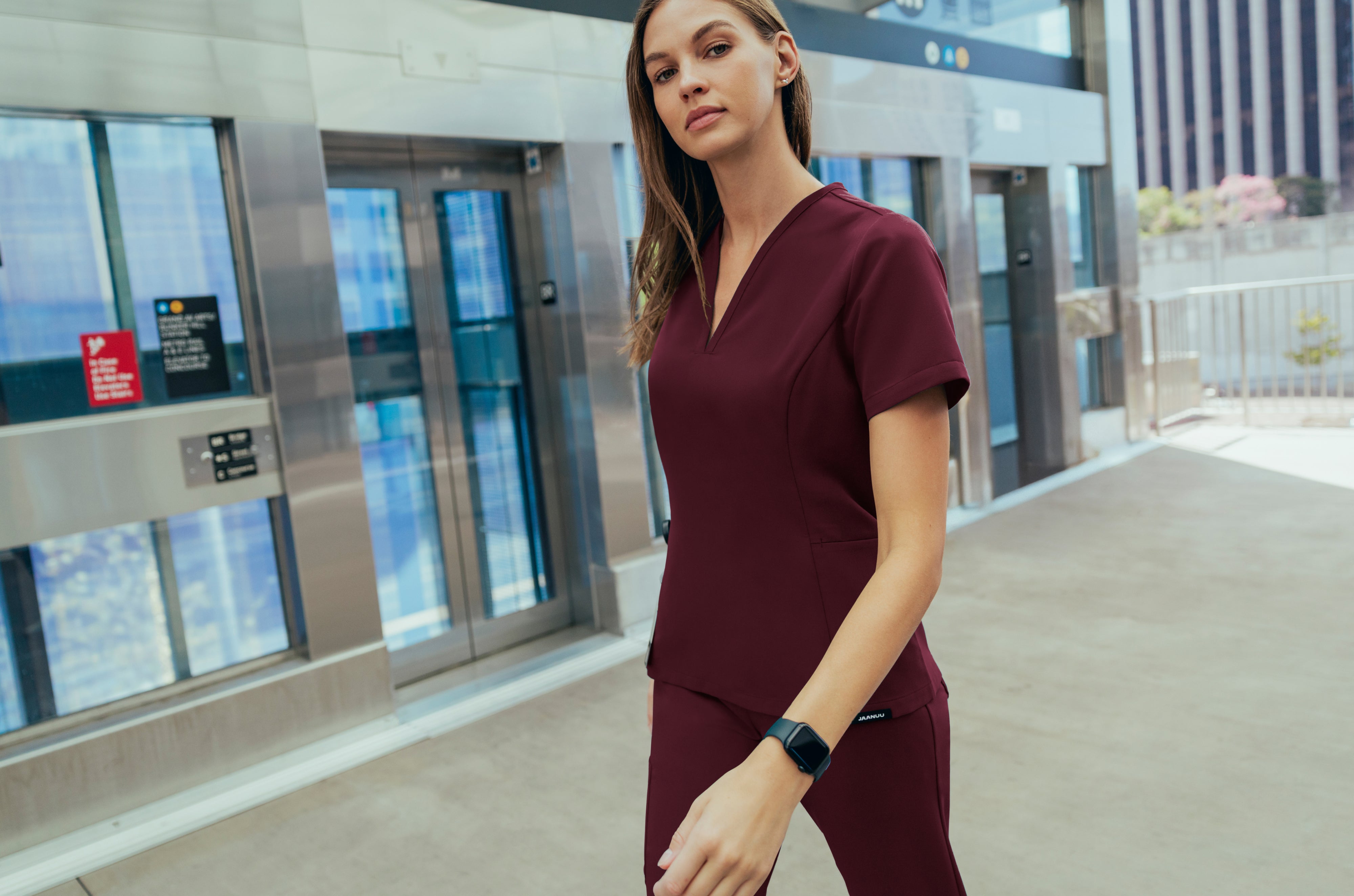 Woman in a dark red Jaanuu V-neck scrub top and scrub pants walking past elevator doors on a city transit platform