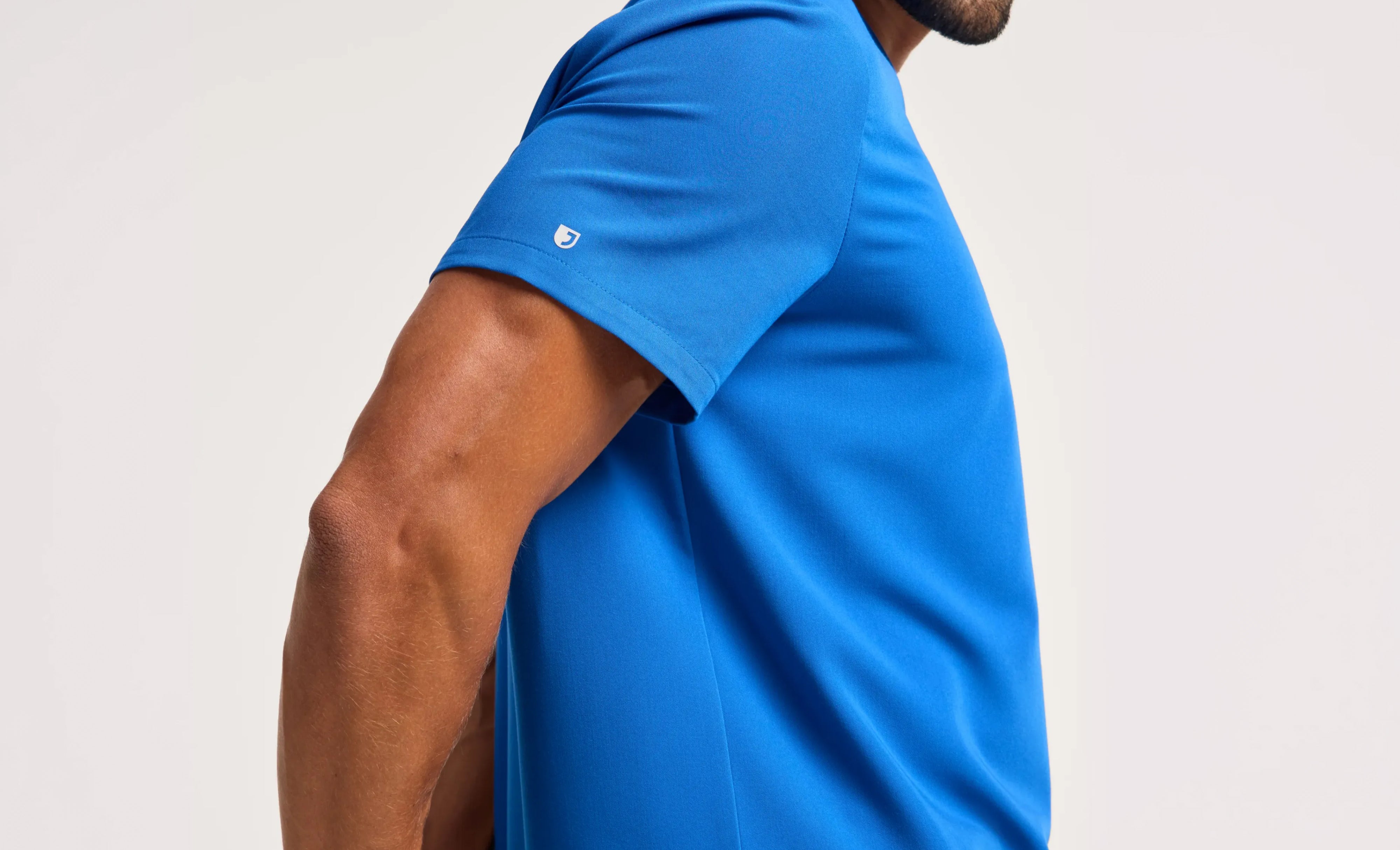Close-up side view of a man's shoulder and arm in a bright blue Jaanuu scrub top with the white Jaanuu J logo on the sleeve, against a light gray backdrop