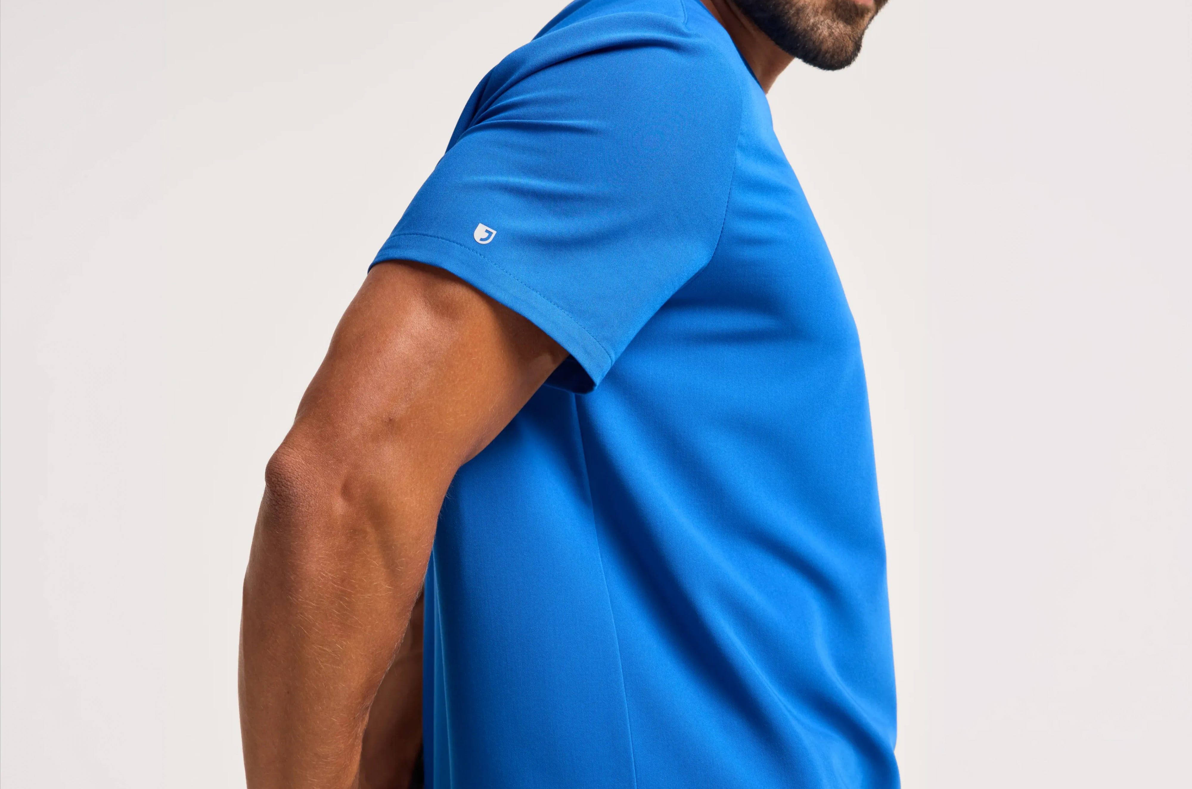 Close-up side view of a man's shoulder and arm in a bright blue Jaanuu scrub top with the white Jaanuu J logo on the sleeve, against a light gray backdrop
