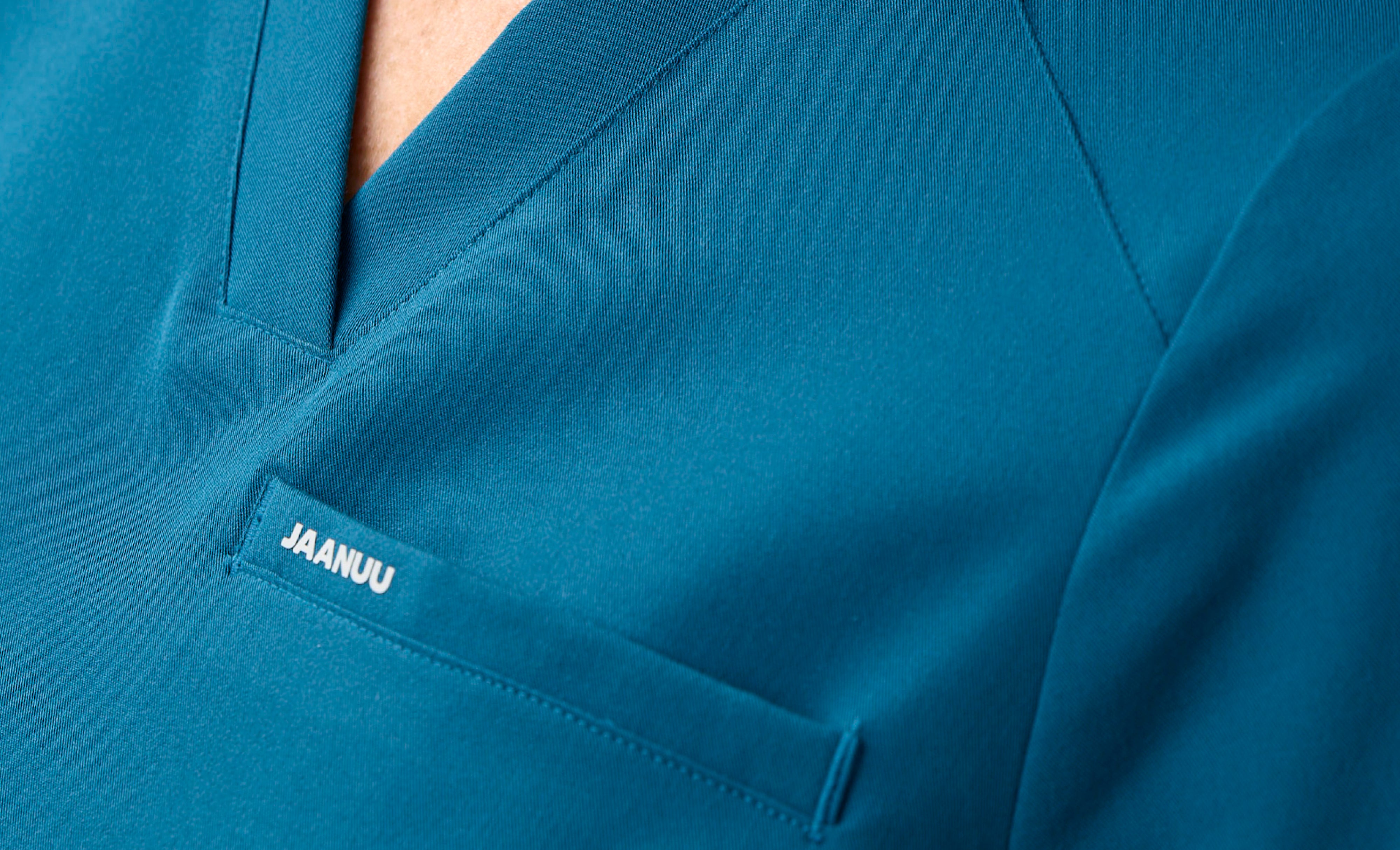 Extreme close-up of the V-neckline and chest pocket of a teal Jaanuu scrub top, with 'JAANUU' printed in white on the pocket