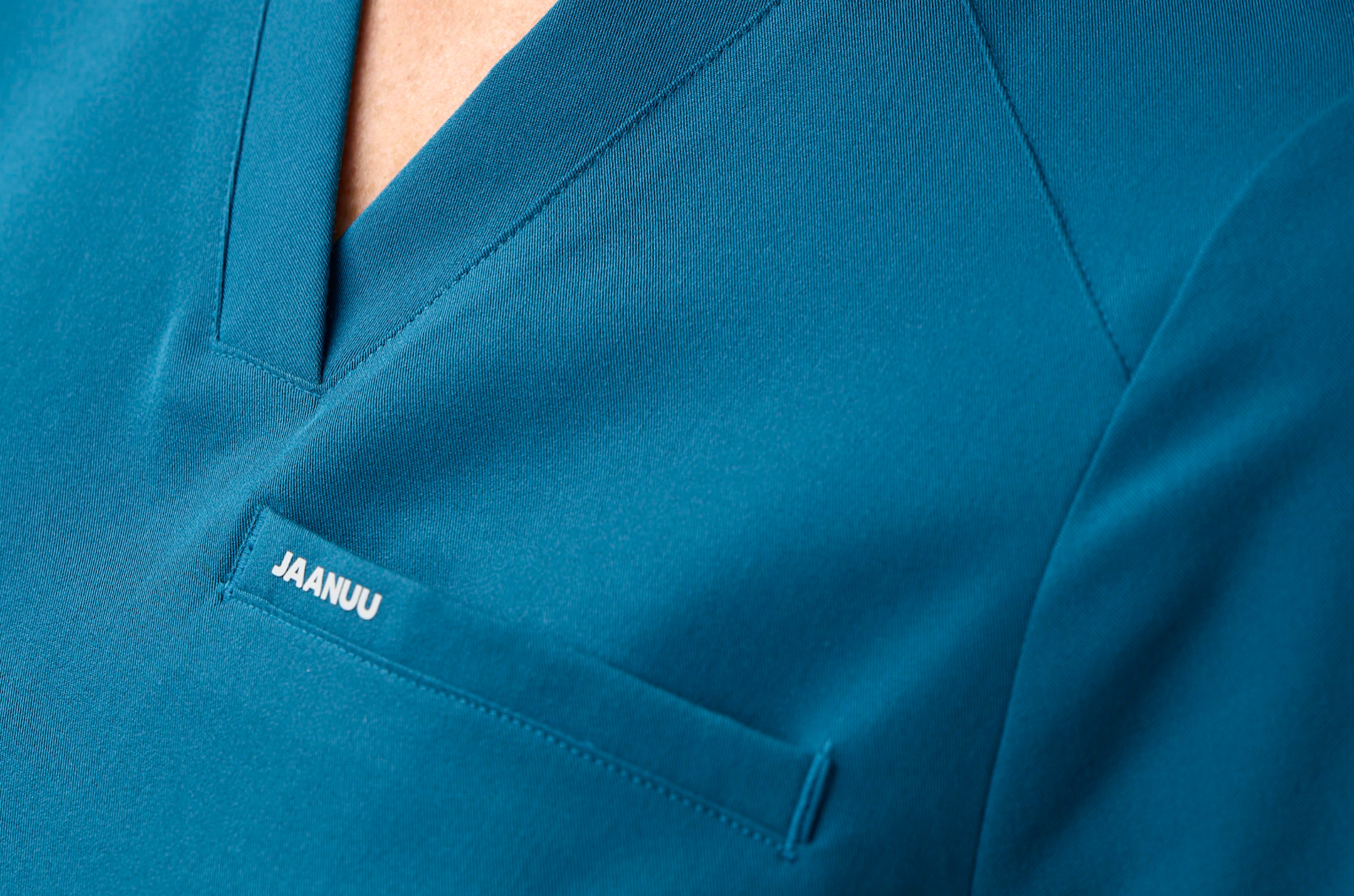 Extreme close-up of the V-neckline and chest pocket of a teal Jaanuu scrub top, with 'JAANUU' printed in white on the pocket
