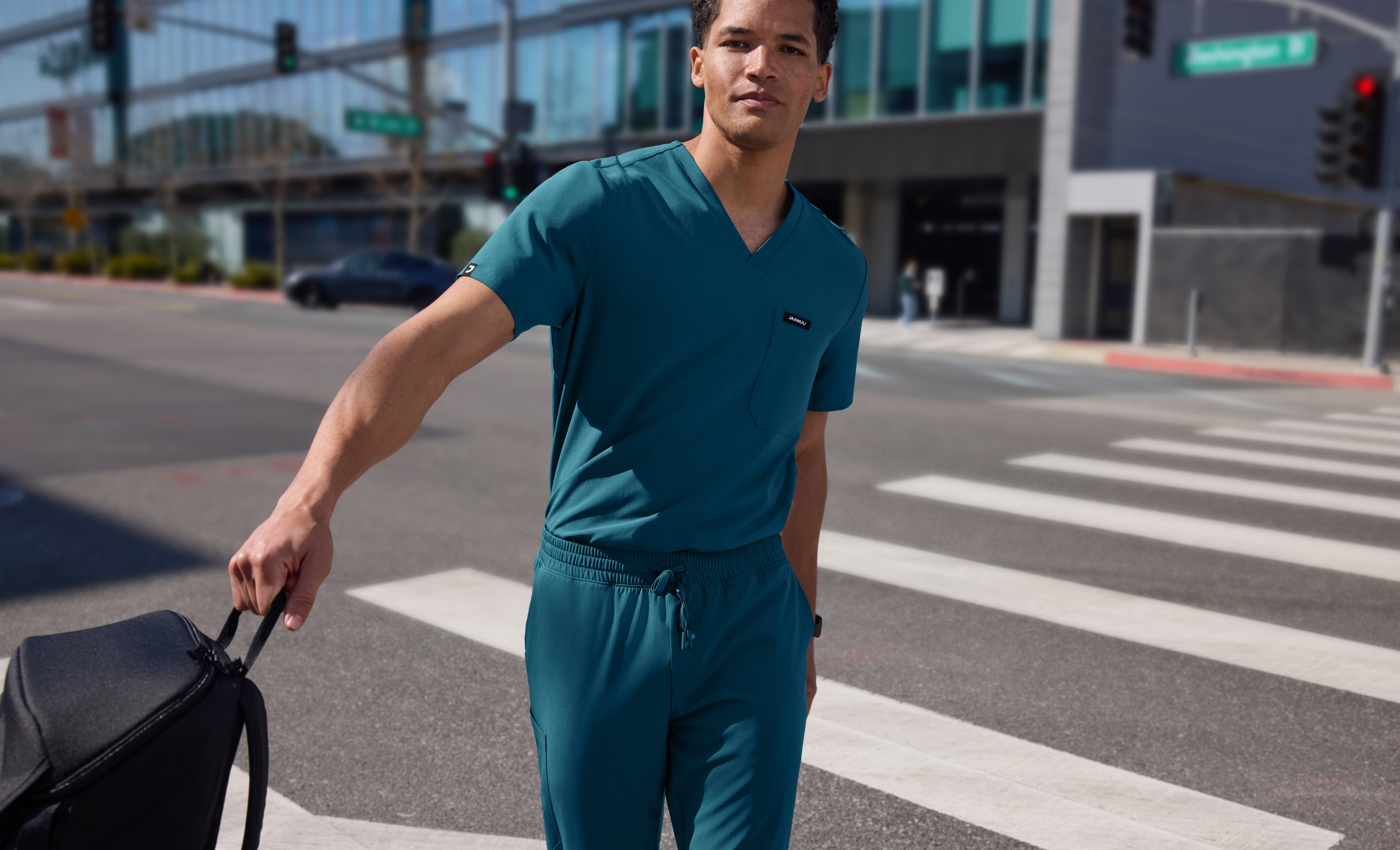 Man in a teal Jaanuu V-neck scrub top and jogger scrub pants carrying a black backpack across a city crosswalk, with glass buildings behind him