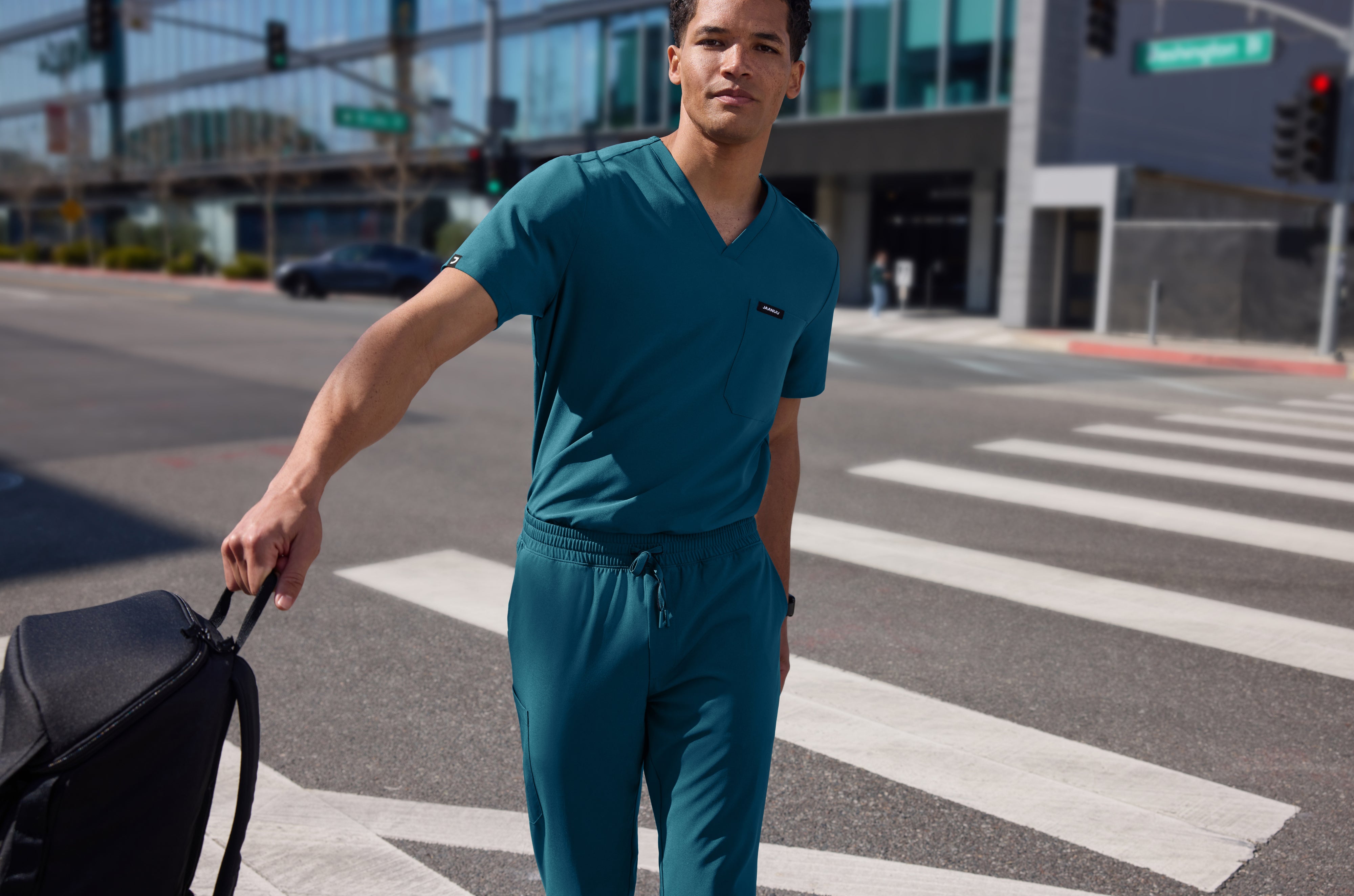 Man in a teal Jaanuu V-neck scrub top and jogger scrub pants carrying a black backpack across a city crosswalk, with glass buildings behind him