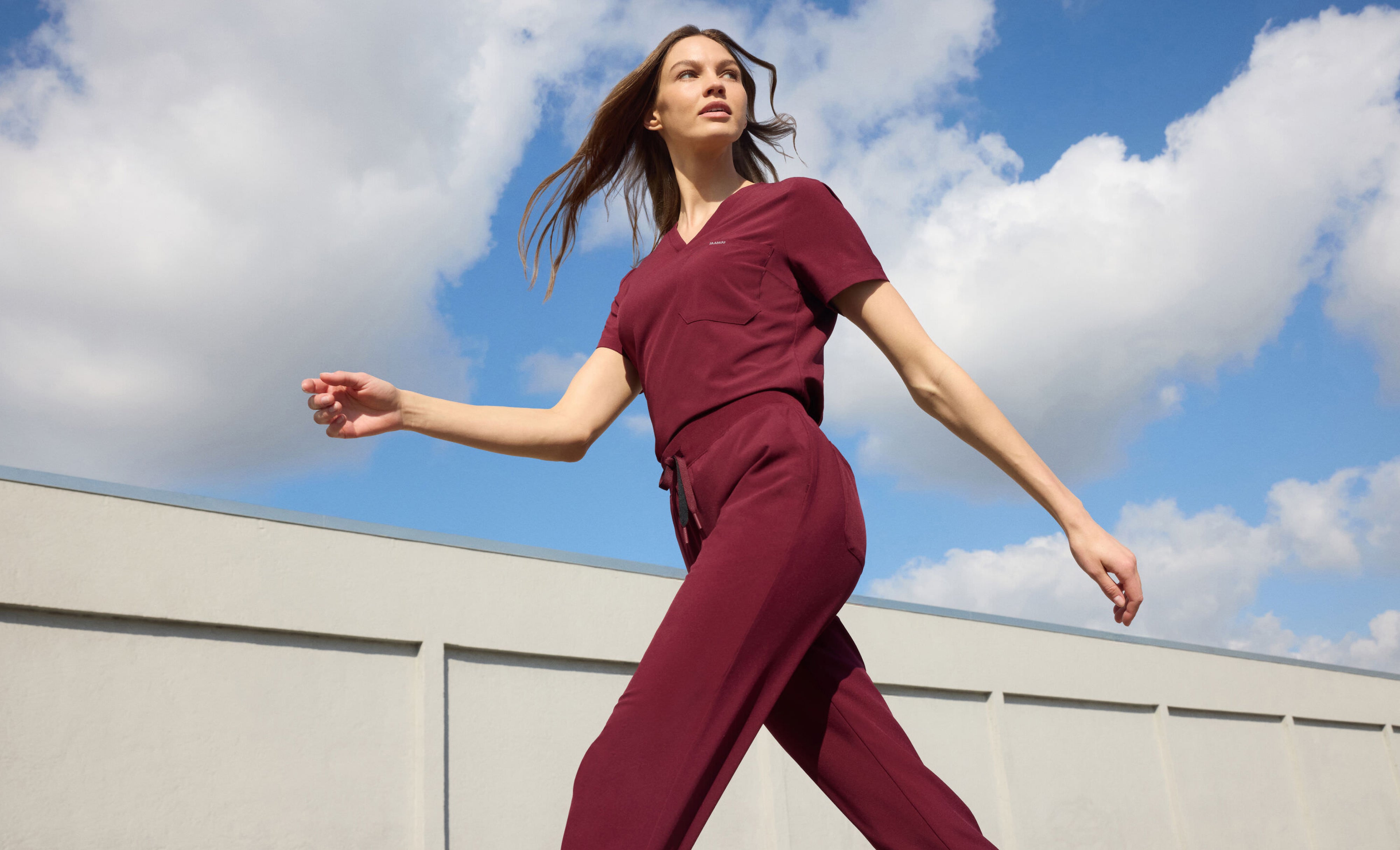 Woman in a dark red Jaanuu V-neck scrub top and drawstring scrub pants striding past a white wall under a blue sky with clouds