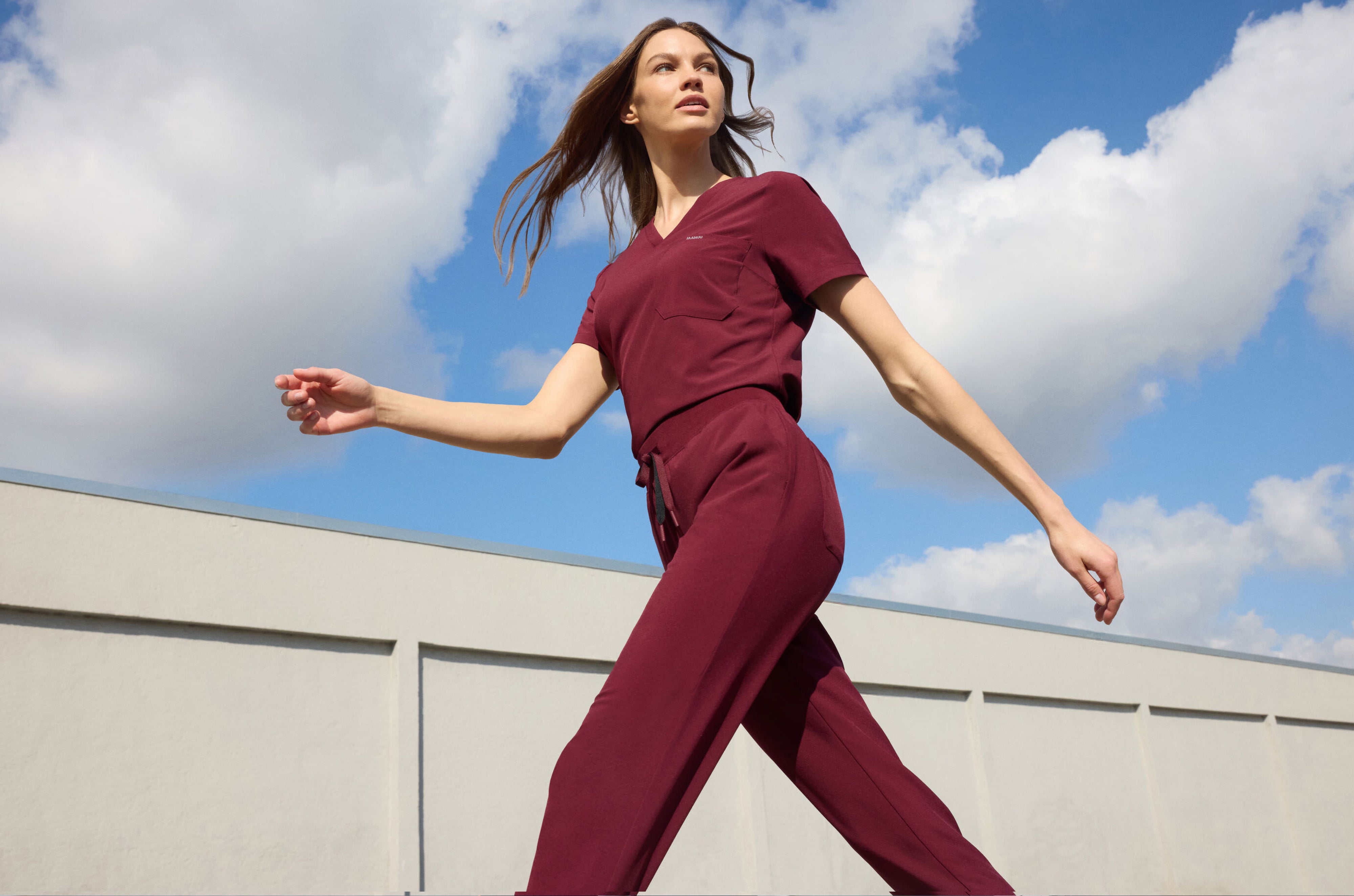Woman in a dark red Jaanuu V-neck scrub top and drawstring scrub pants striding past a white wall under a blue sky with clouds