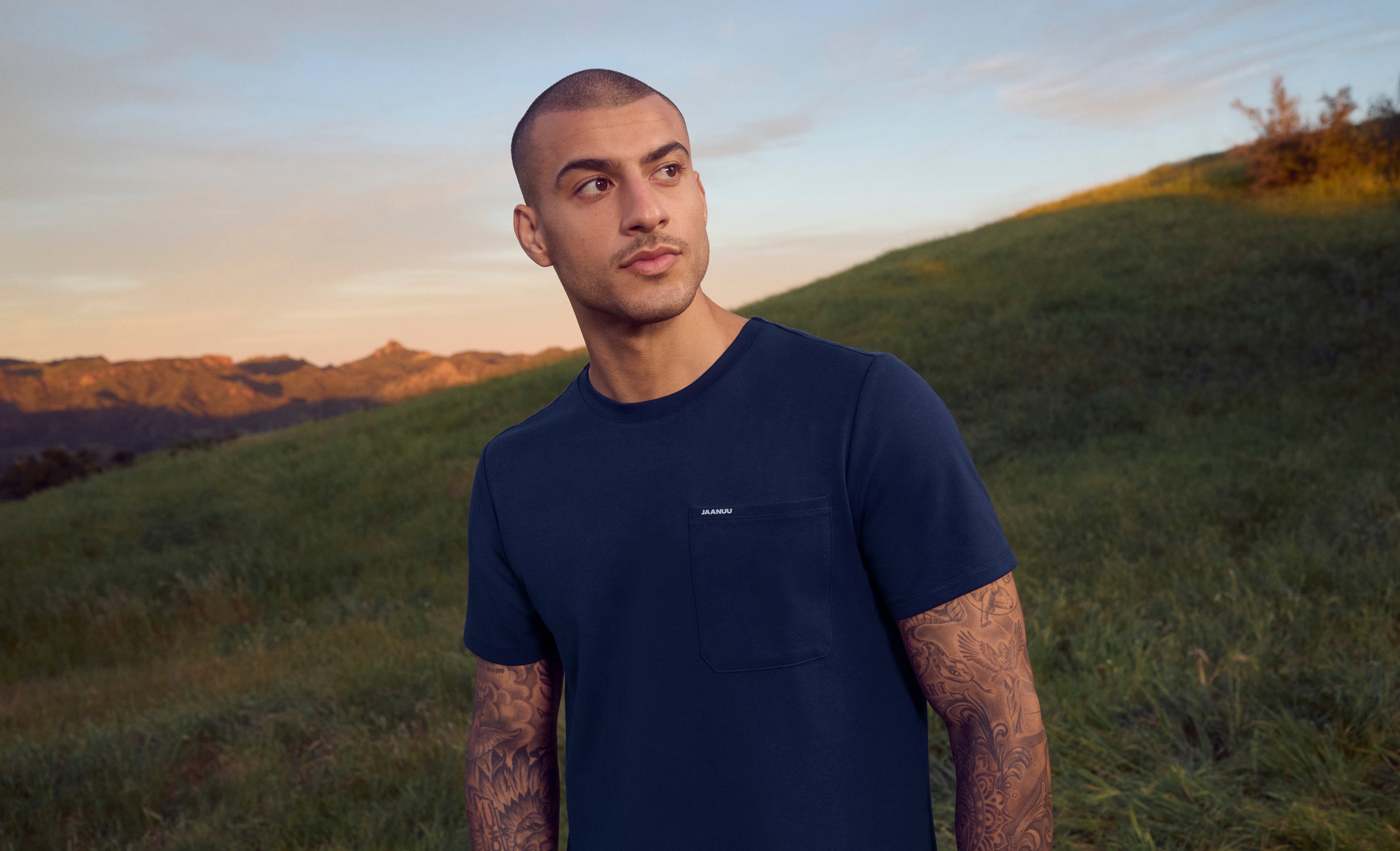 Man in a navy blue Jaanuu crewneck pocket tee standing on a grassy hillside at sunset, with mountains behind him